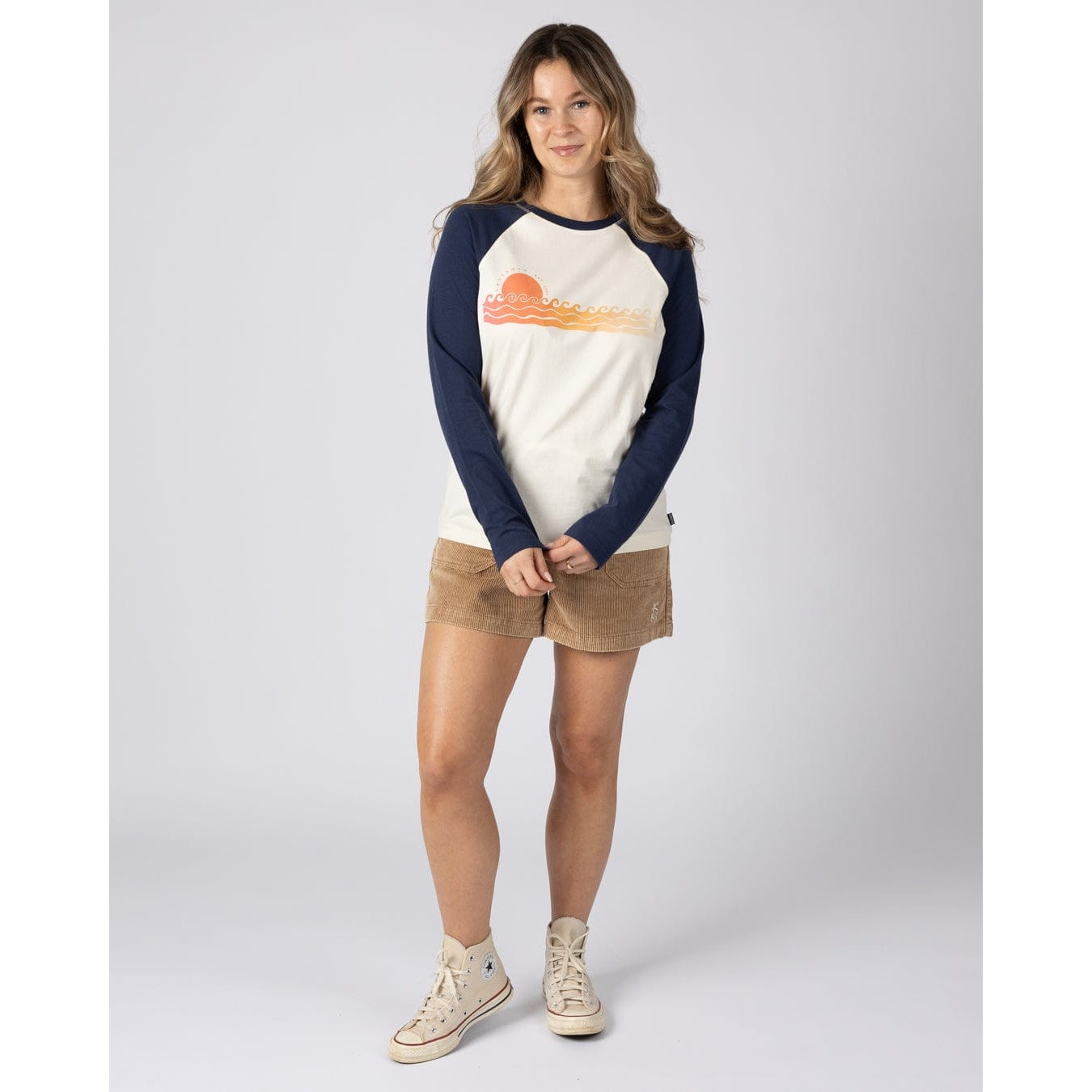 Woman wearing a Saltrock womens Golden Hour raglan t-shirt in cream and navy with tan colour shorts and cream sneakers on a plain background