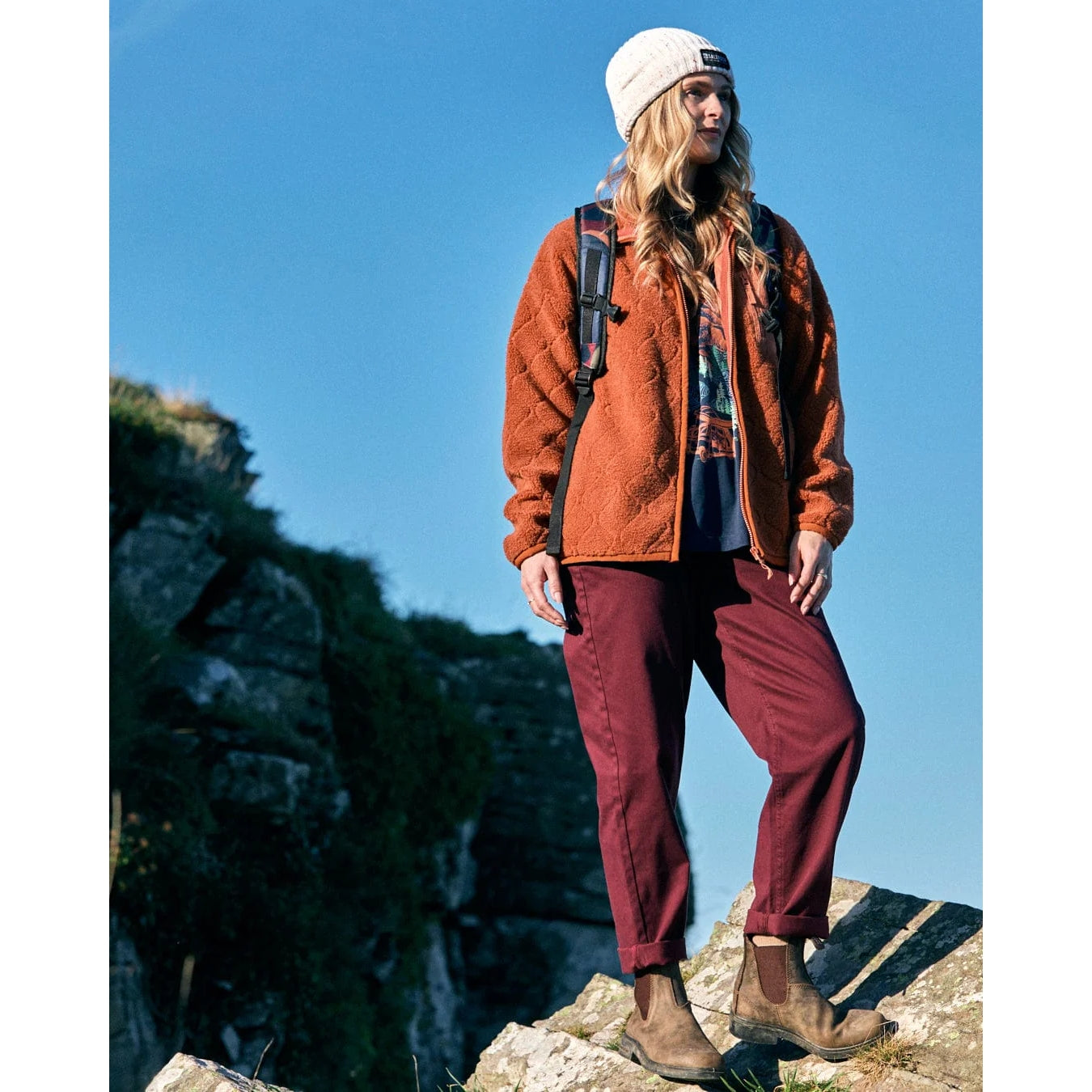 Woman wearing an orange fleece jacket and maroon pants standing on a rocky outcrop with a clear blue sky.