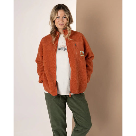 Woman wearing an orange Saltrock Jarvis fleece, white shirt, and green pants against a beige background