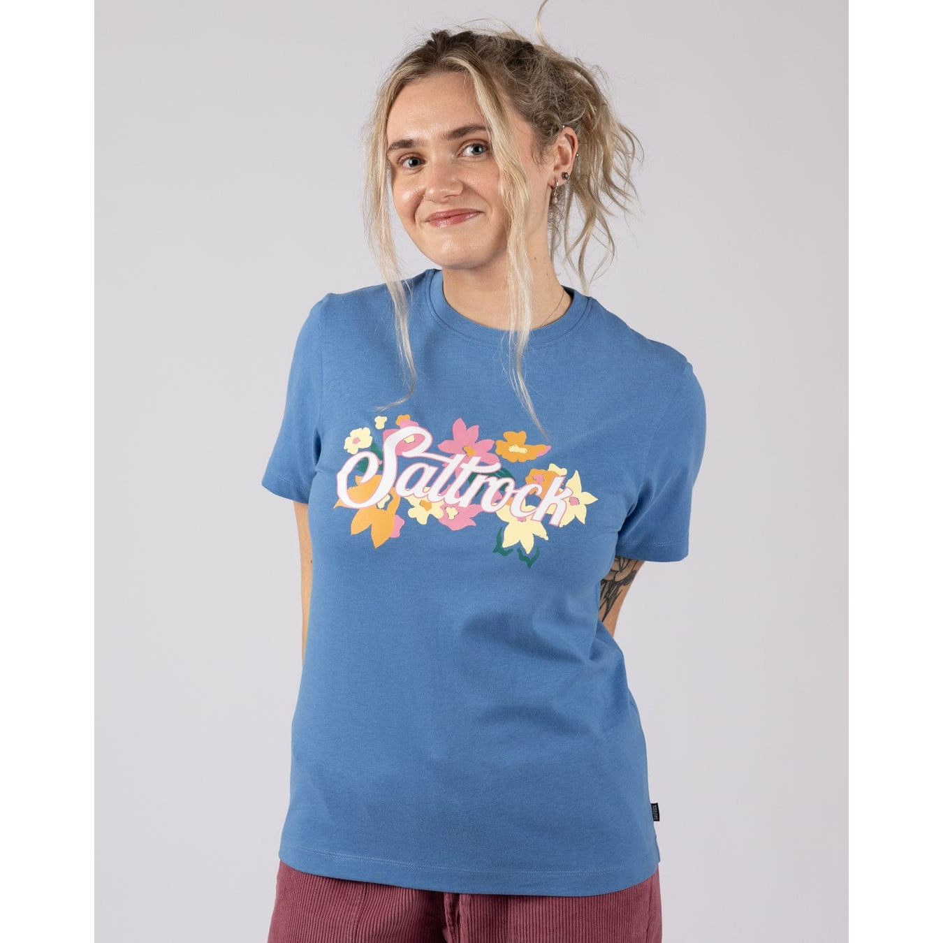 Front view of a woman wearing a Saltrock short sleeve Trader Daze t-shirt in bright blue with a floral logo print on the chest with burgundy trousers on a plain background