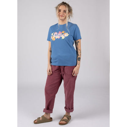 A woman wearing a blue Saltrock short sleeve Trader Daze tee with floral logo print, burgundy trousers and sandals against a plain background