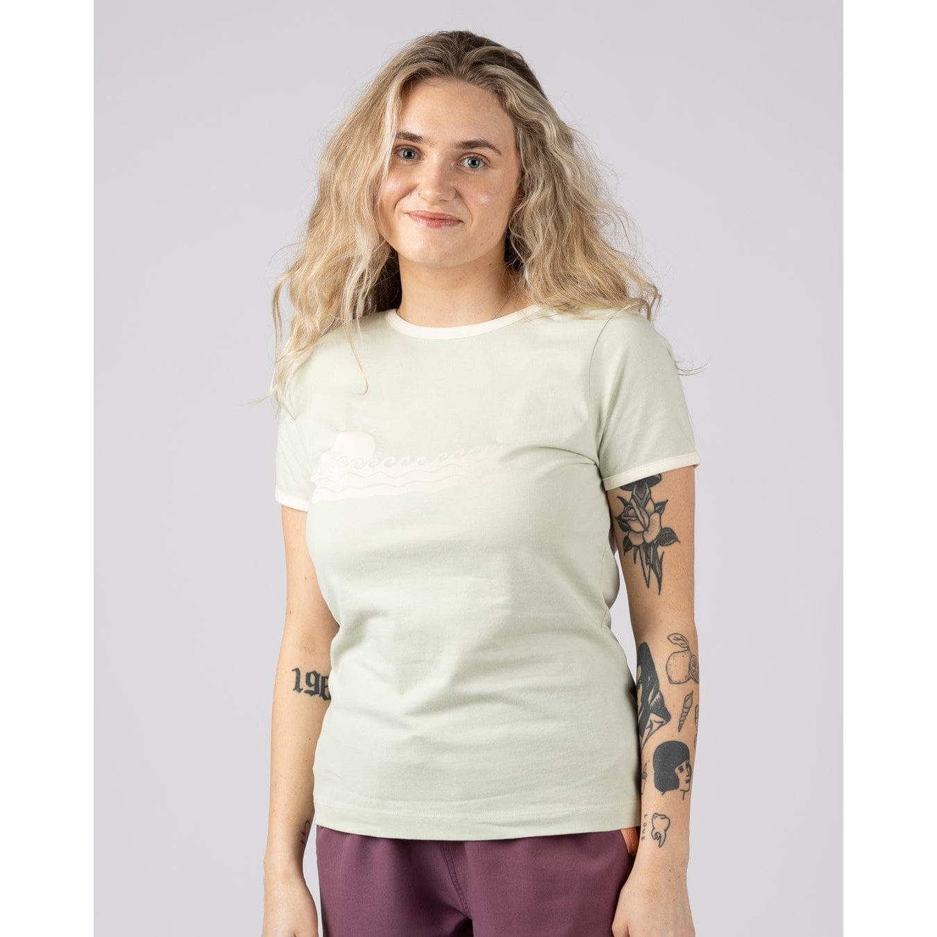 Front view of a woman wearing a light green Saltrock Golden Hour short sleeve t-shirt with sunset and sea print across the chest with burgundy shorts against a plain background