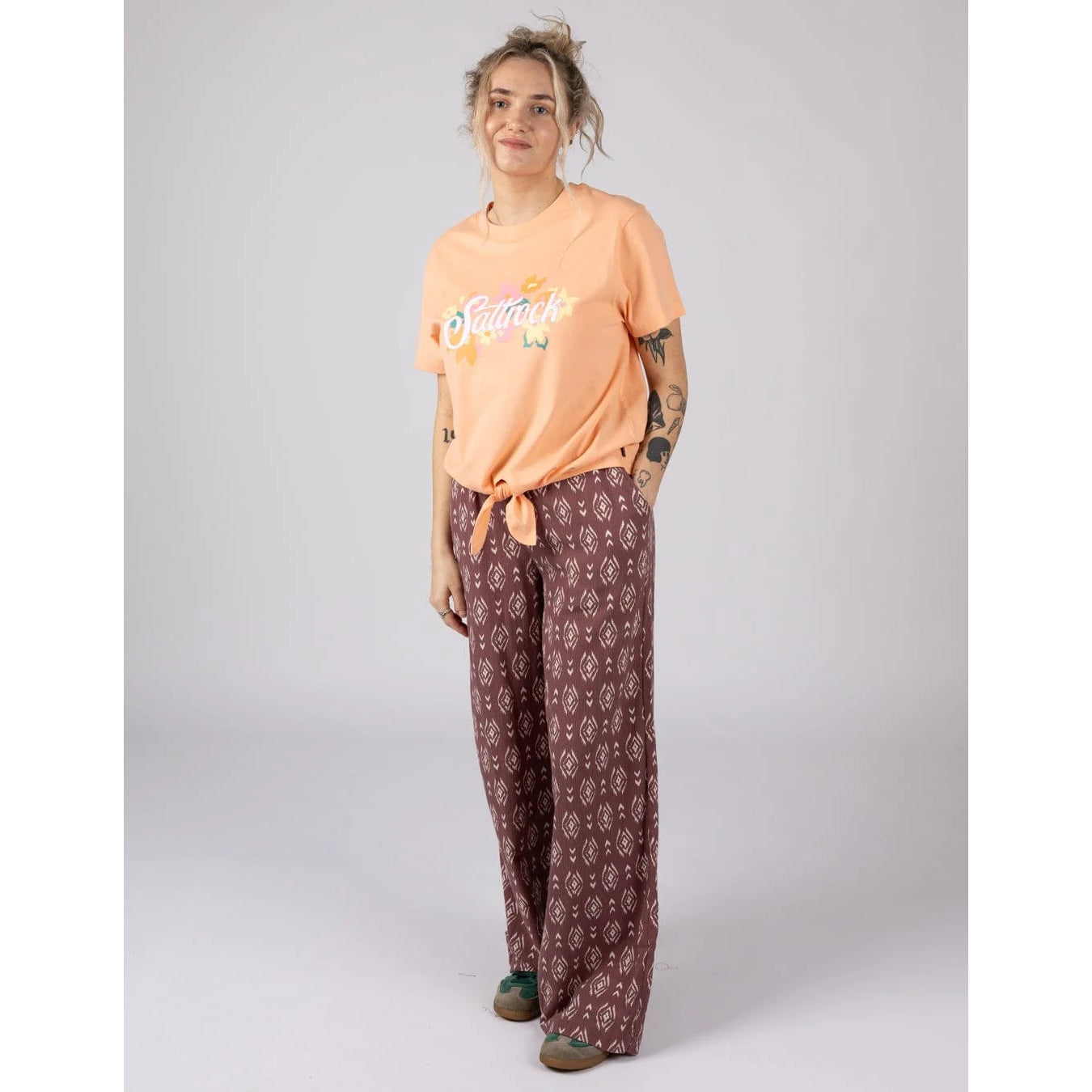 Woman wearing a Saltrock short sleeve Trader Daze t-shirt in peach, with logo print on the chest and a knot tie hem with burgundy trousers.