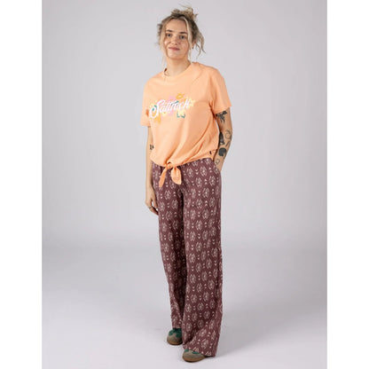 Woman wearing a Saltrock short sleeve Trader Daze t-shirt in peach, with logo print on the chest and a knot tie hem with burgundy trousers.