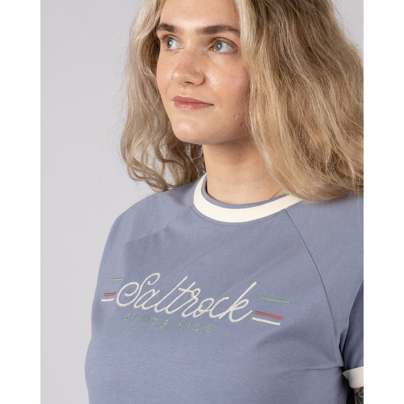 Close-up view of a woman wearing a blue short sleeve Saltrock Links t-shirt with embroidered chest logo on a plain background