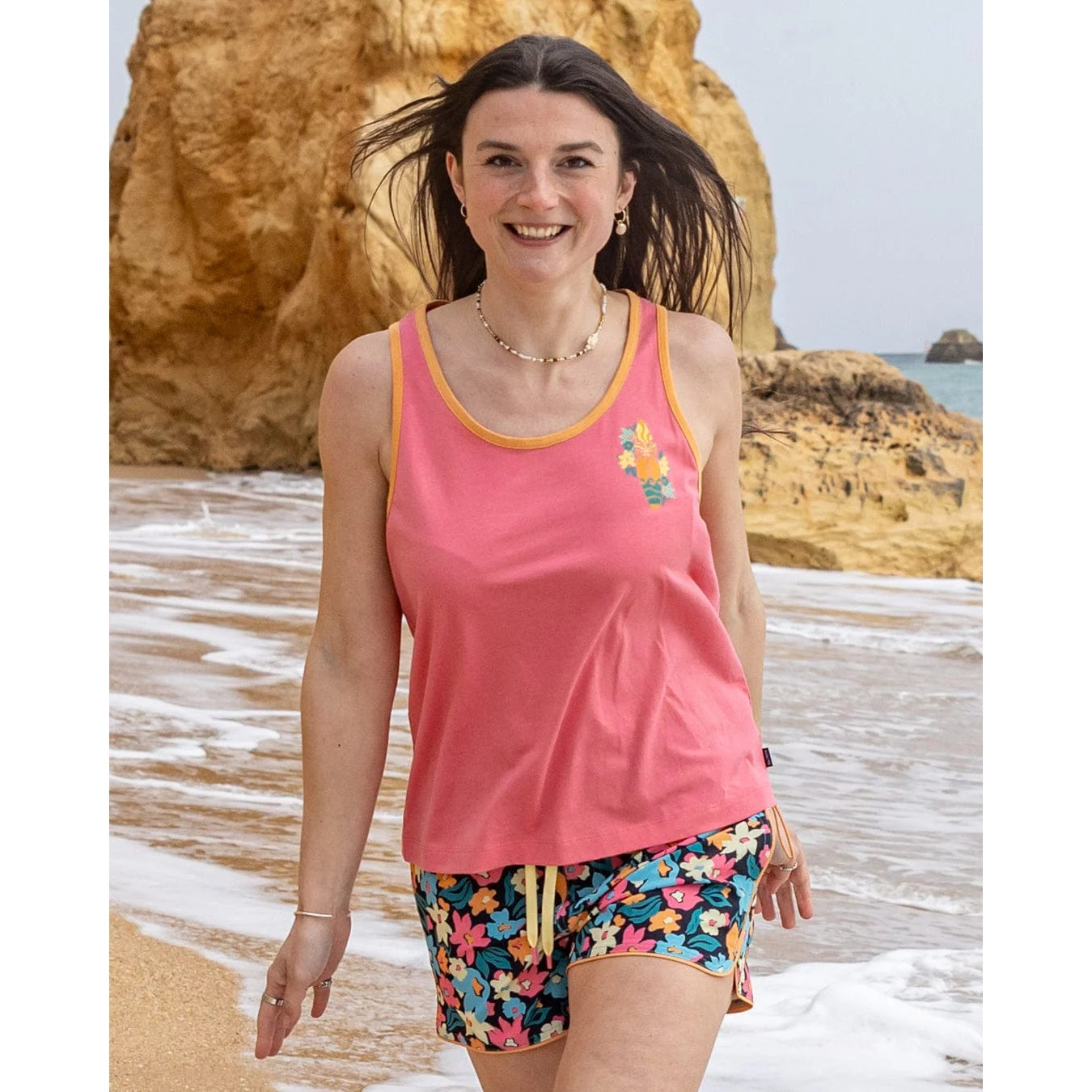 Woman wearing a pink Saltrock Sun Surfer vest top and colourful shorts standing on a beach with ocean and rocks in the background.