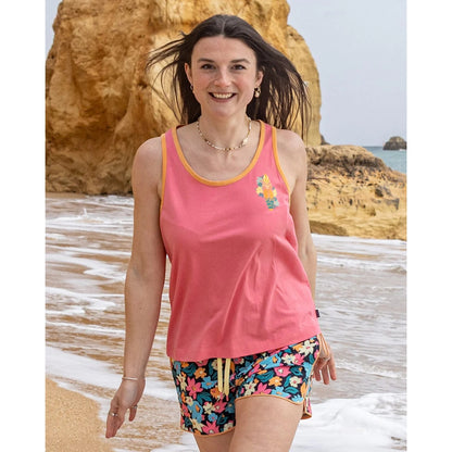Woman wearing a pink Saltrock Sun Surfer vest top and colourful shorts standing on a beach with ocean and rocks in the background.