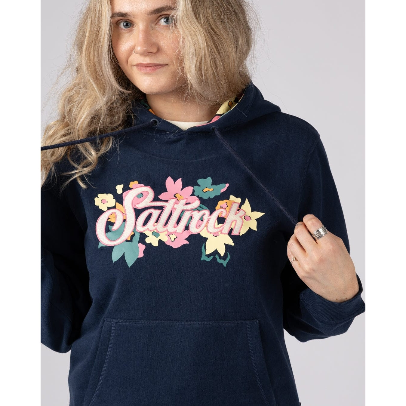 Woman wearing a navy Trader Daze hoodie with colourful 'Saltrock' text and floral designs on a plain background