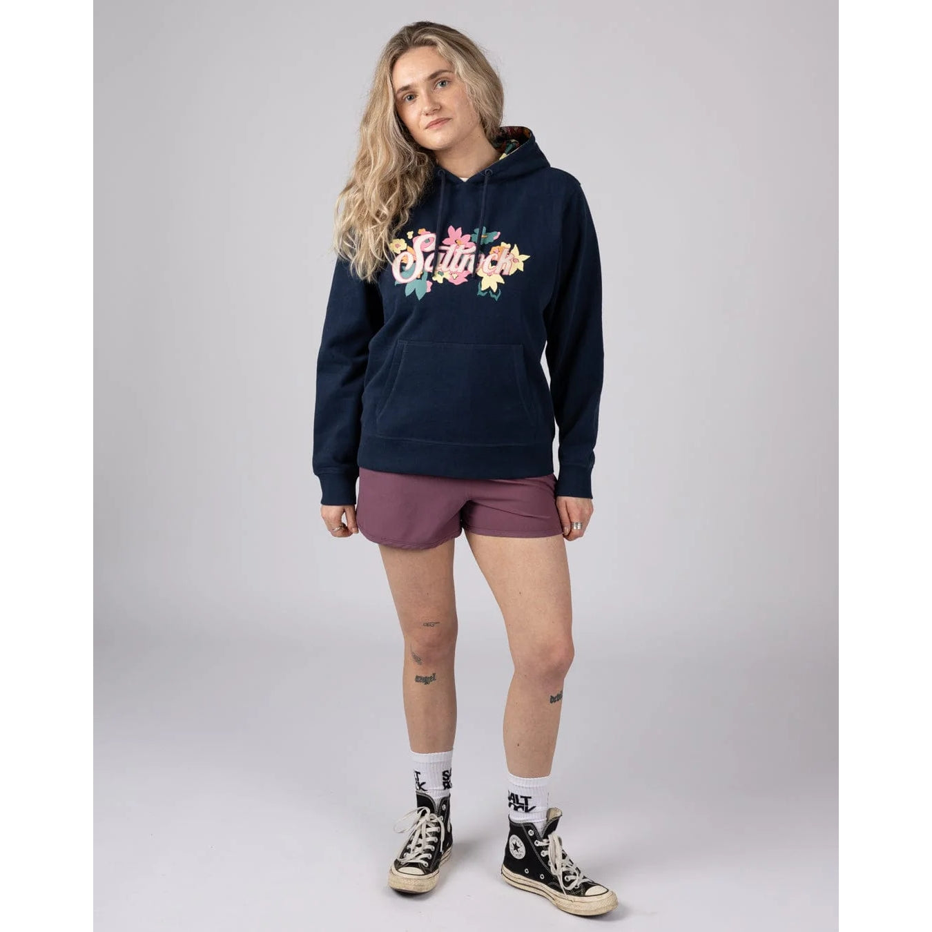 Woman wearing a navy Saltrock Trader Daze pop hoodie with a floral logo print and maroon shorts on a grey background