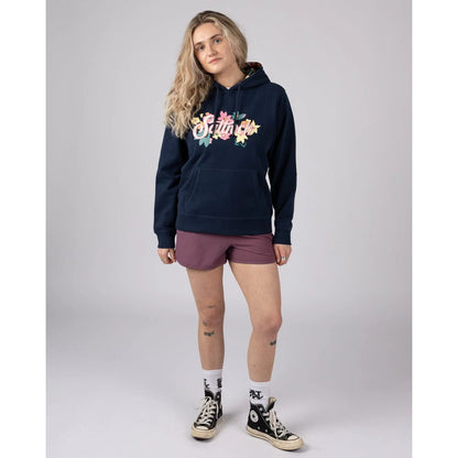 Woman wearing a navy Saltrock Trader Daze pop hoodie with a floral logo print and maroon shorts on a grey background