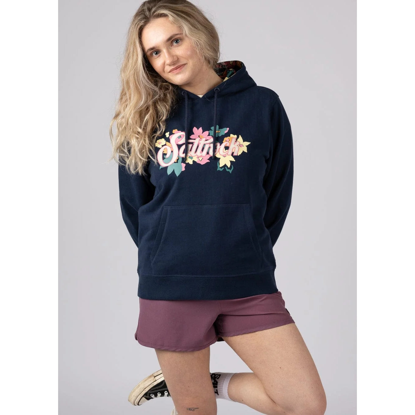 Front view of a woman wearing a navy Saltrock pop hoodie with floral logo design on a plain background