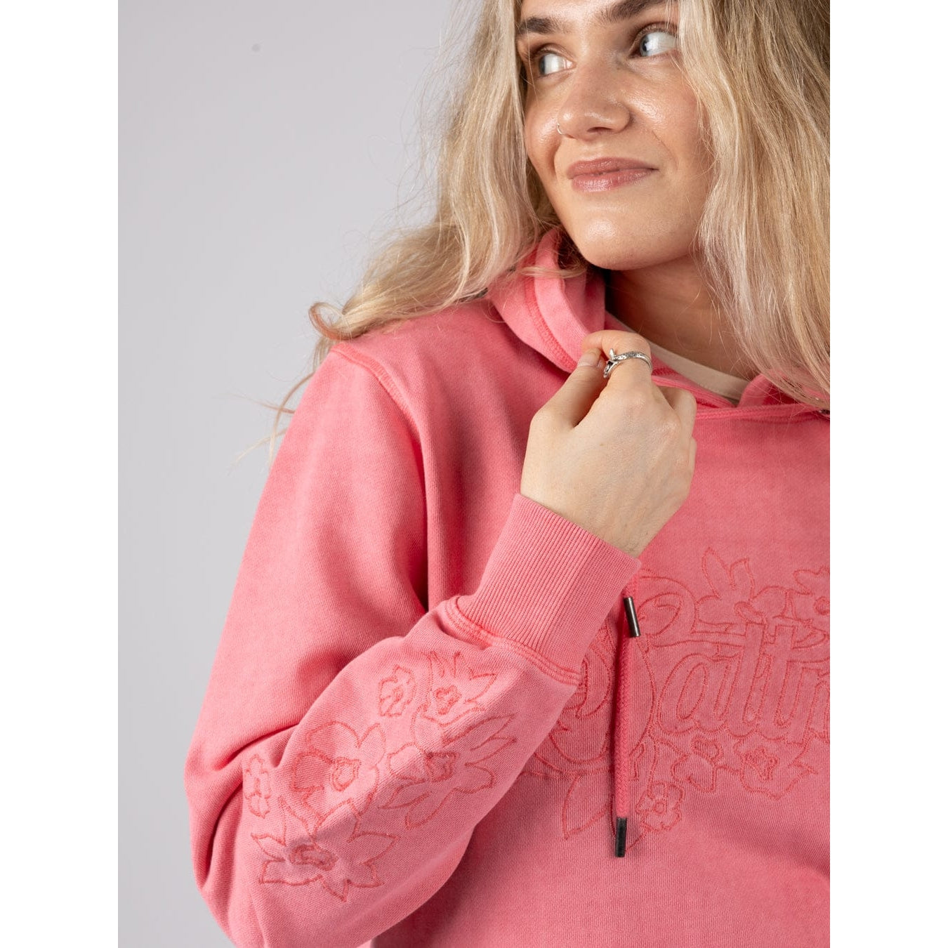 Close-up view of a woman wearing a bright pink Saltrock Trader Tonal hoodie with floral sleeve embroidery on a plain background