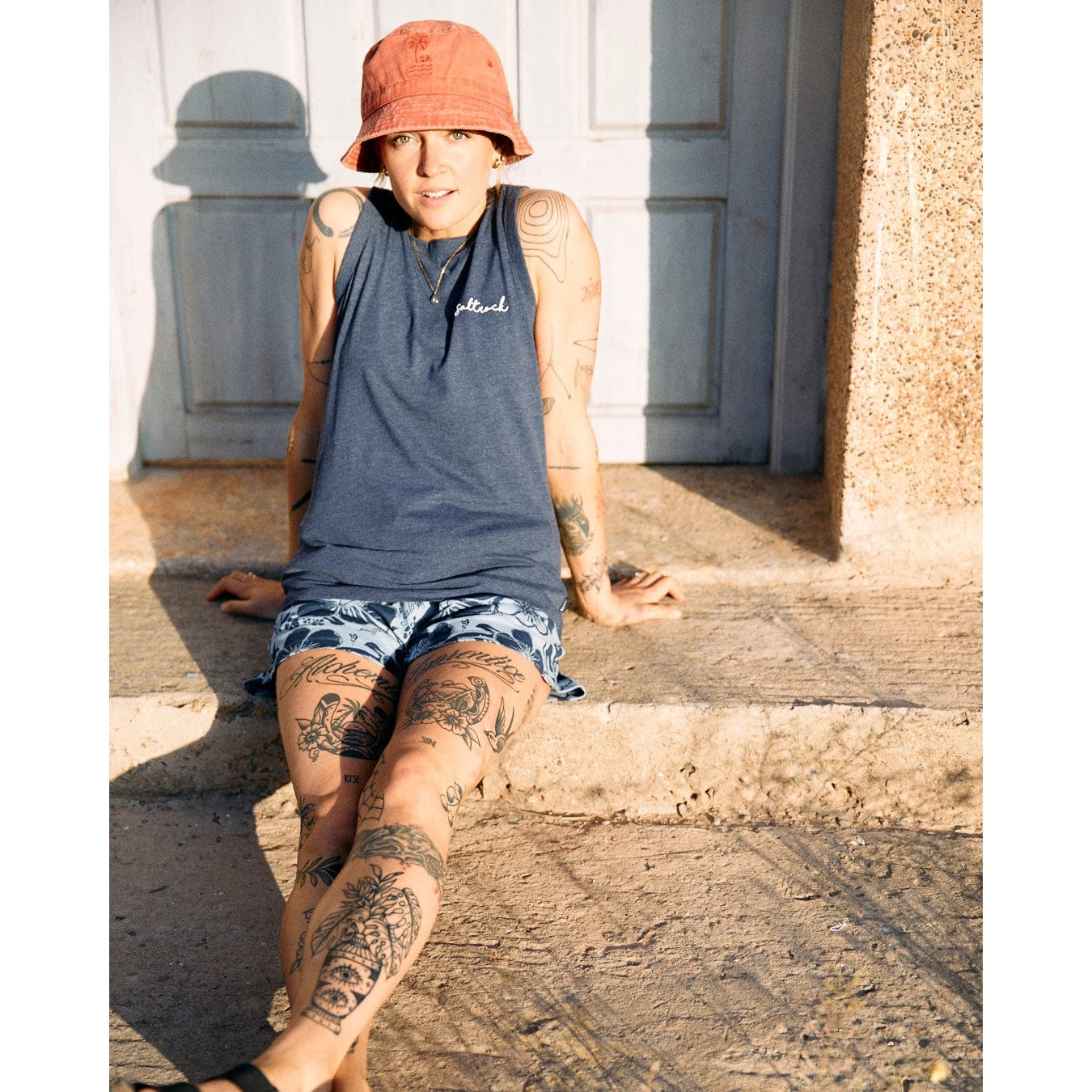 A woman wearing a blue marl sleeveless Saltrock Velator vest with blue floral shorts sat on a step outside