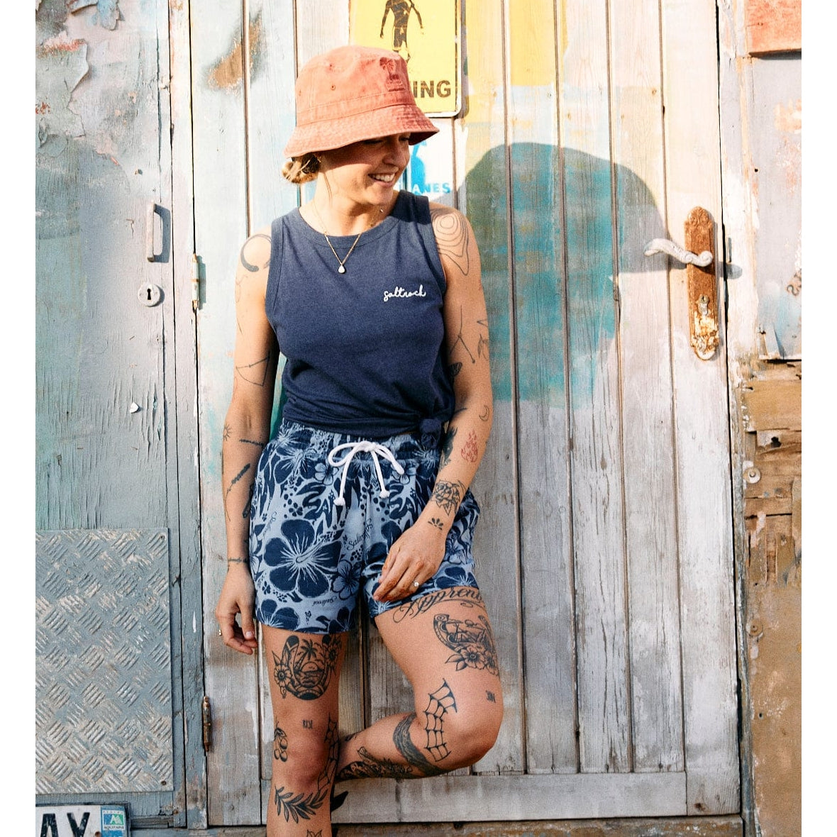 A woman wearing a blue marl Saltrock Velator vest with blue floral shorts stood outside again a door