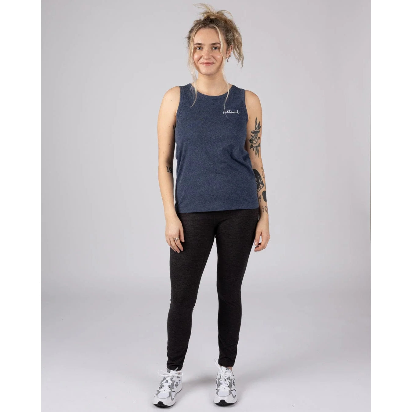 Woman wearing a a blue marl Saltrock Velator vest with black trousers and white trainers on a plain background