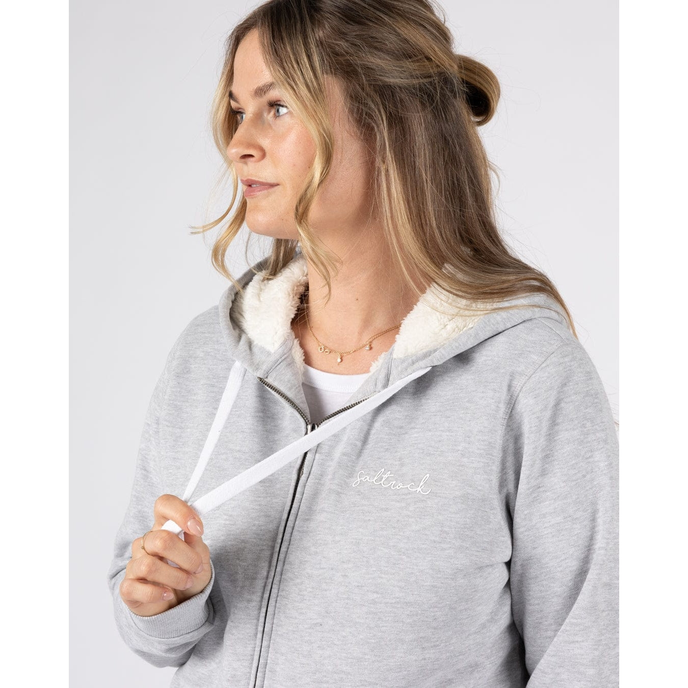 Close-up of a woman wearing a grey Saltrock full zip Velator hoodie with white fur lining on a plain background