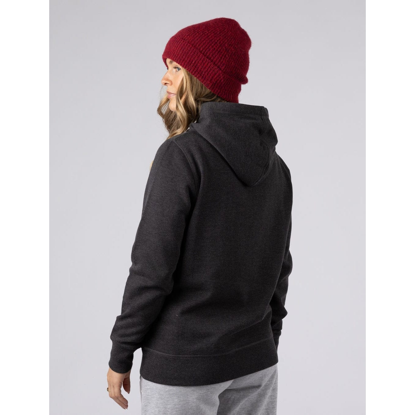 Back view of a woman wearing a dark grey Saltrock pop hoodie and red beanie on a grey background