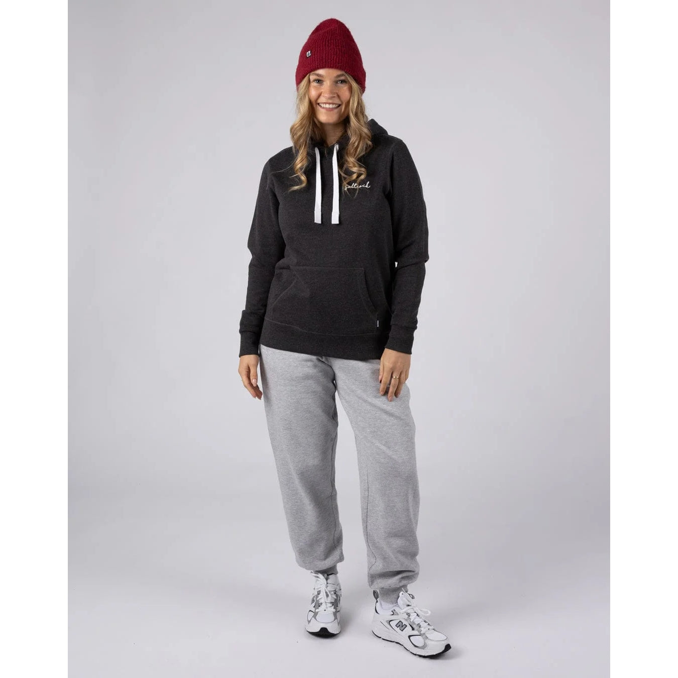 Woman wearing a dark grey Saltrock pop hoodie, grey joggers, and a red beanie on a light grey background