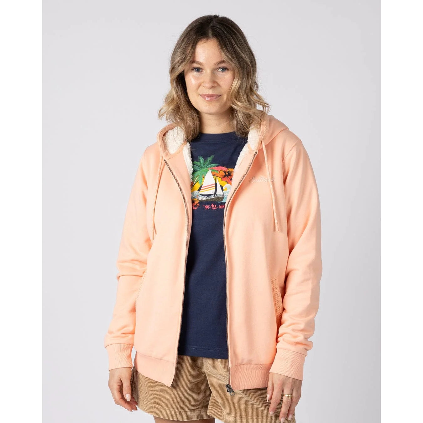 Front view of a woman wearing a peach or light pink Saltrock Tamsin hoodie with borg lining over a navy graphic t-shirt on a white background