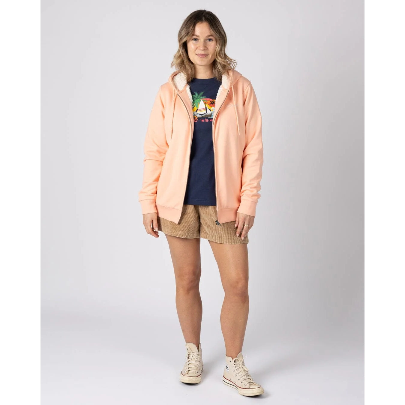 Woman wearing a peach or light pink Tamsin fur lined hoodie, navy t-shirt, beige shorts, and white sneakers on a light grey background