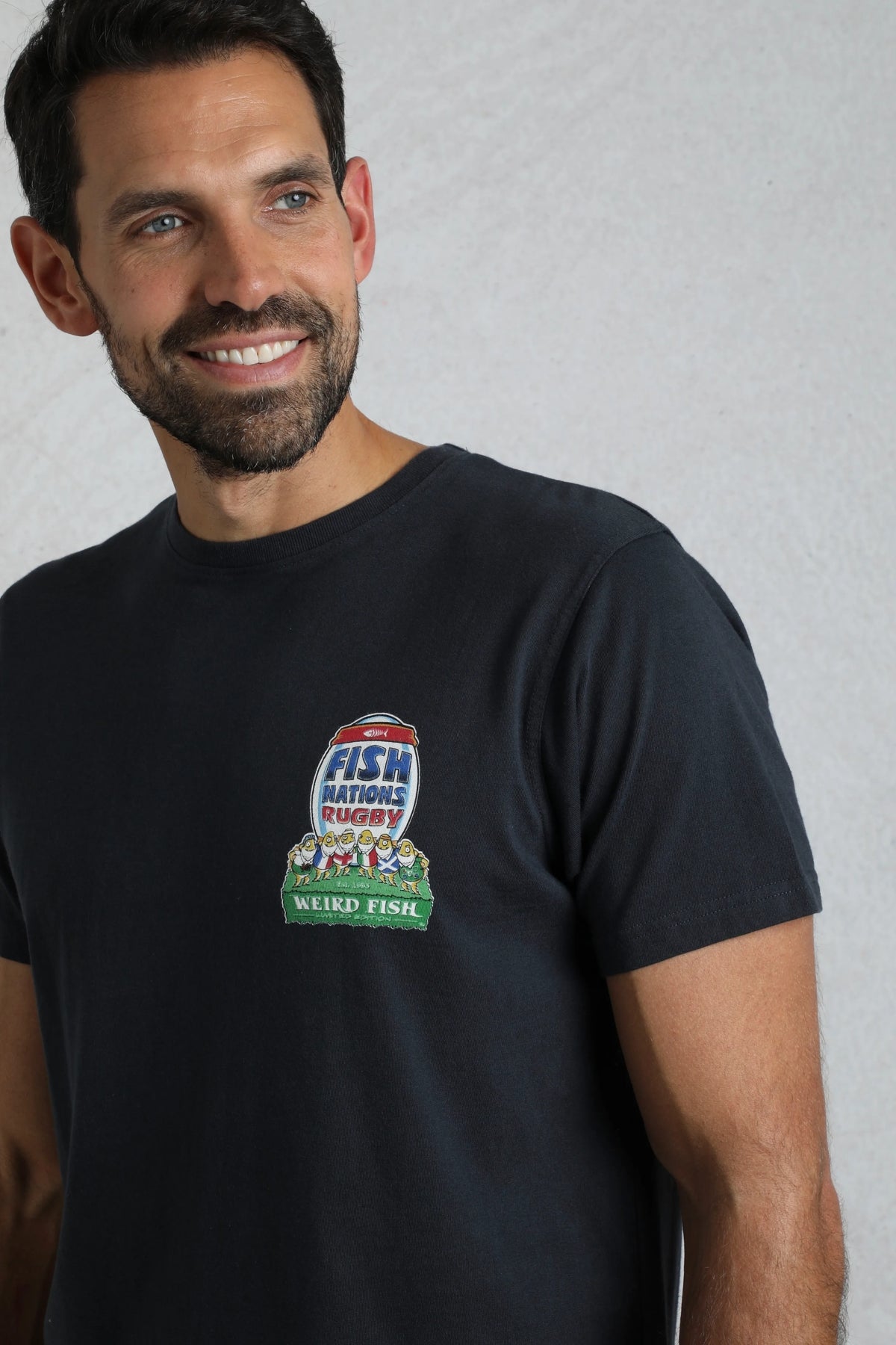 Man wearing a Weird Fish navy short sleeve t-shirt with a rugby themed 'Fish Nations' print on the front, on a white background