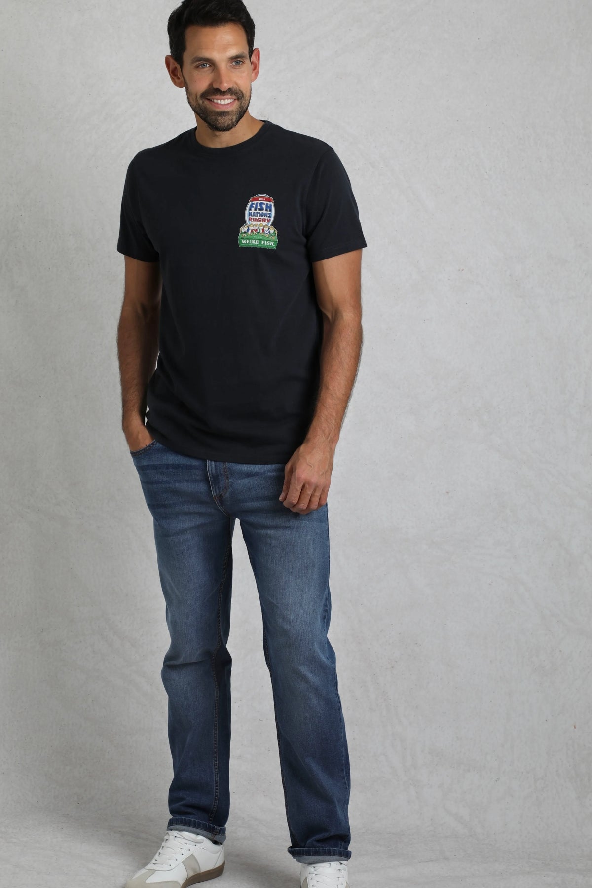 Man wearing a navy Weird Fish short sleeve Fish Nations rugby themed printed t-shirt with blue jeans on a light grey background.