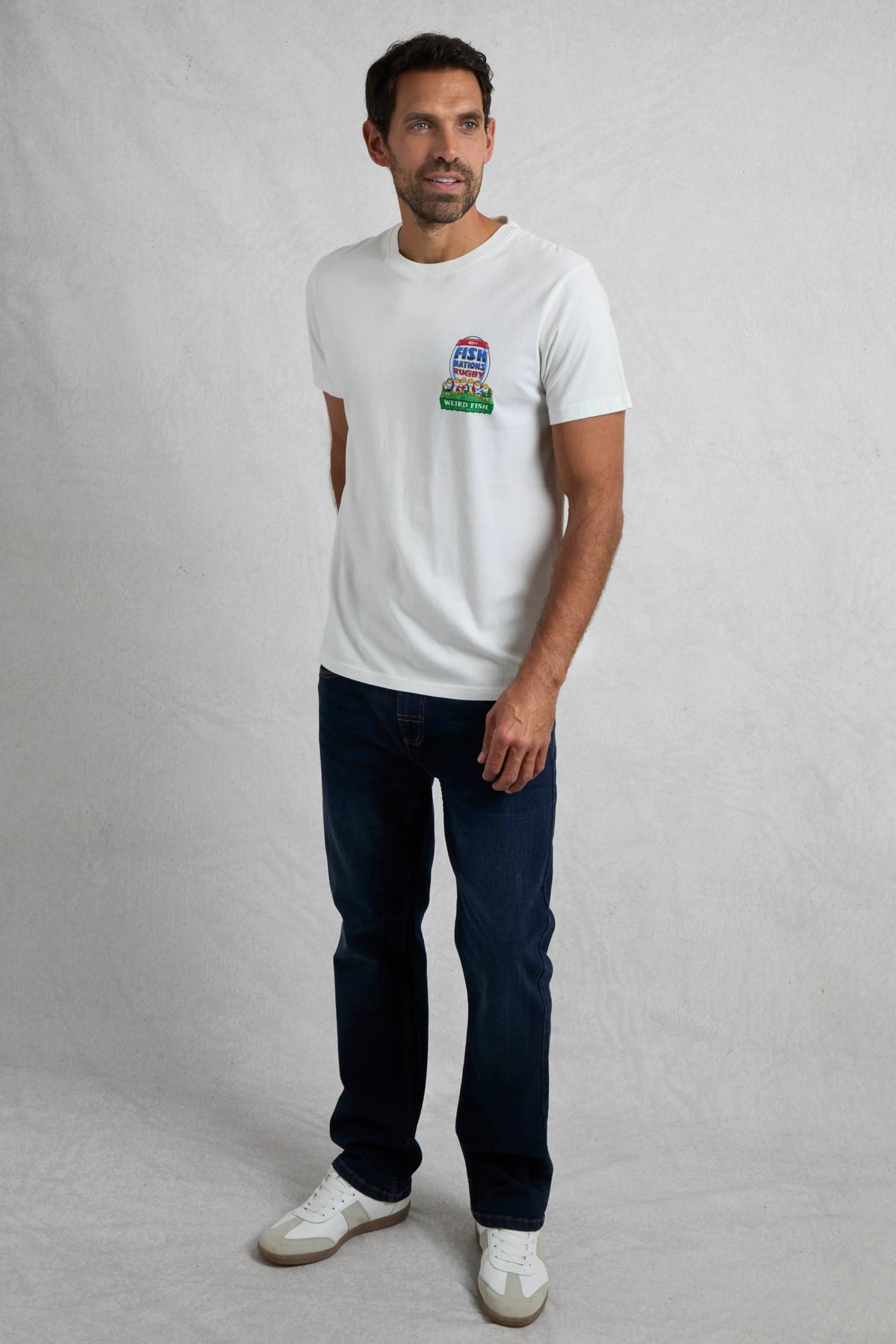 Man wearing a Weird Fish short sleeve white t-shirt with a rugby themed print, with jeans on a white background