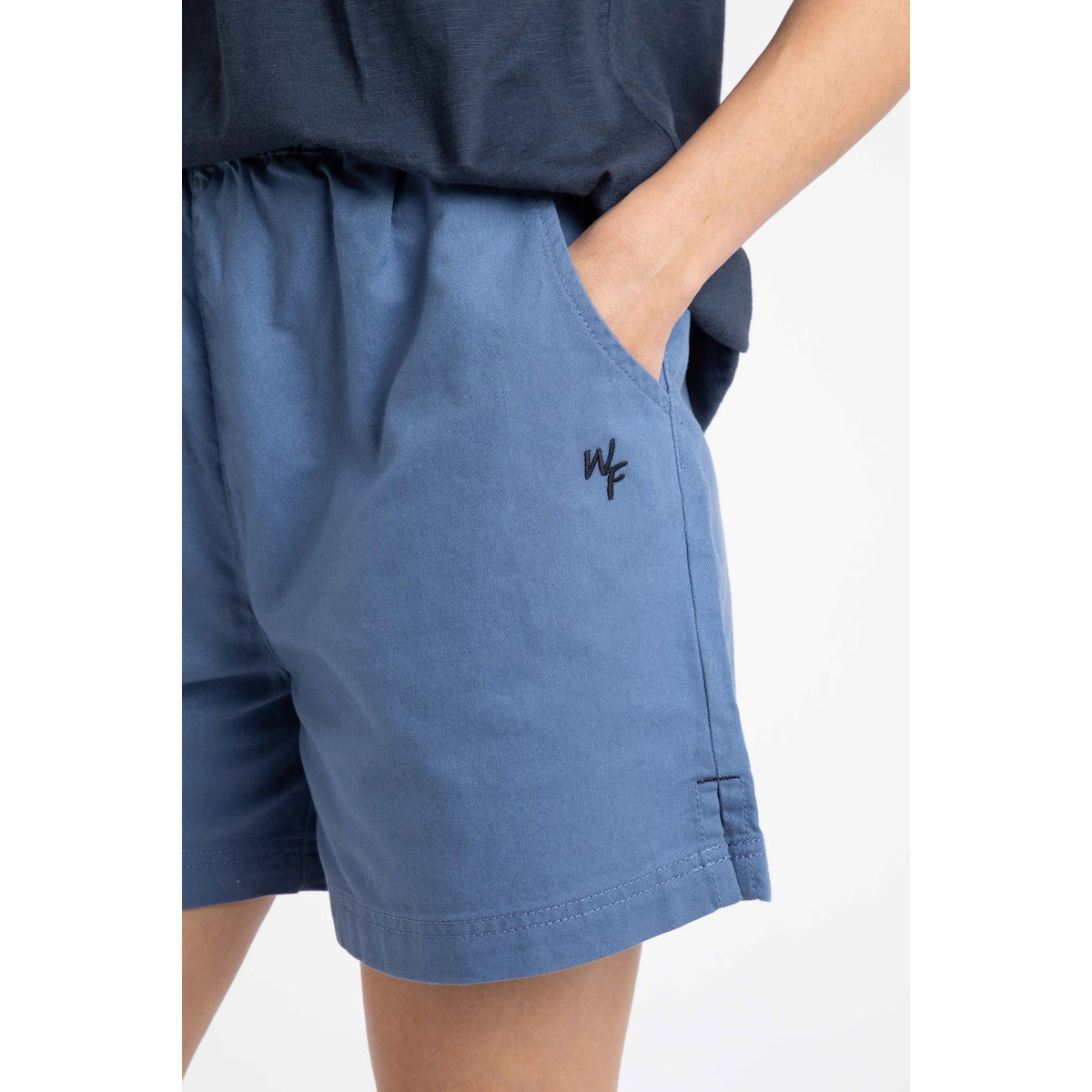 Close-up view of a woman wearing a pair of Weird Fish Jardin elastic waist twill cotton shorts in Faded Blue with hip pockets and small embroidered logo.