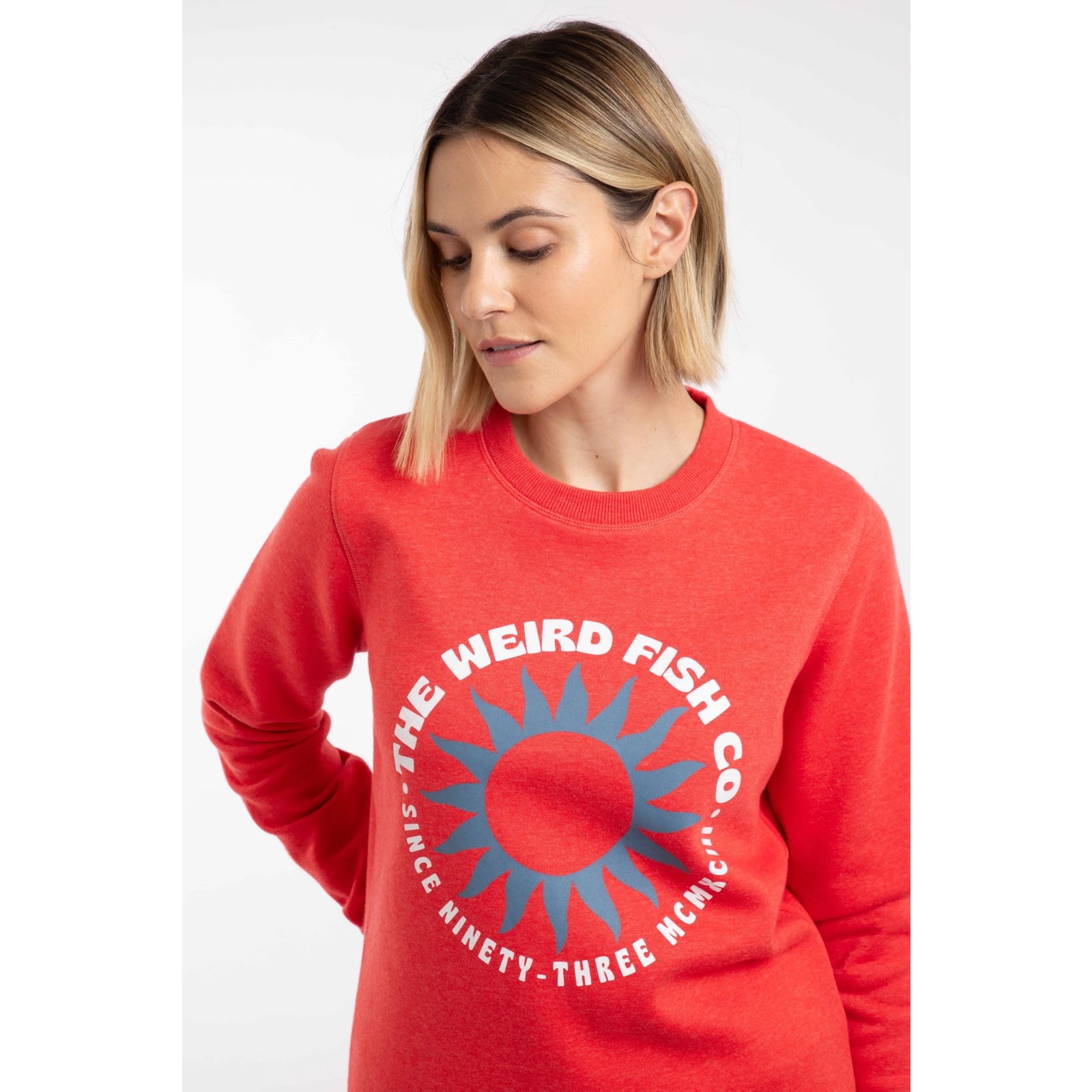 Close-up view of a woman wearing a Weird Fish Aurora crew neck sweatshirt in a bright coral colour with bold circular logo print on the front.