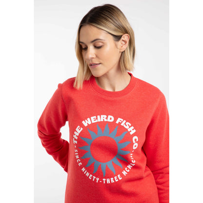 Close-up view of a woman wearing a Weird Fish Aurora crew neck sweatshirt in a bright coral colour with bold circular logo print on the front.