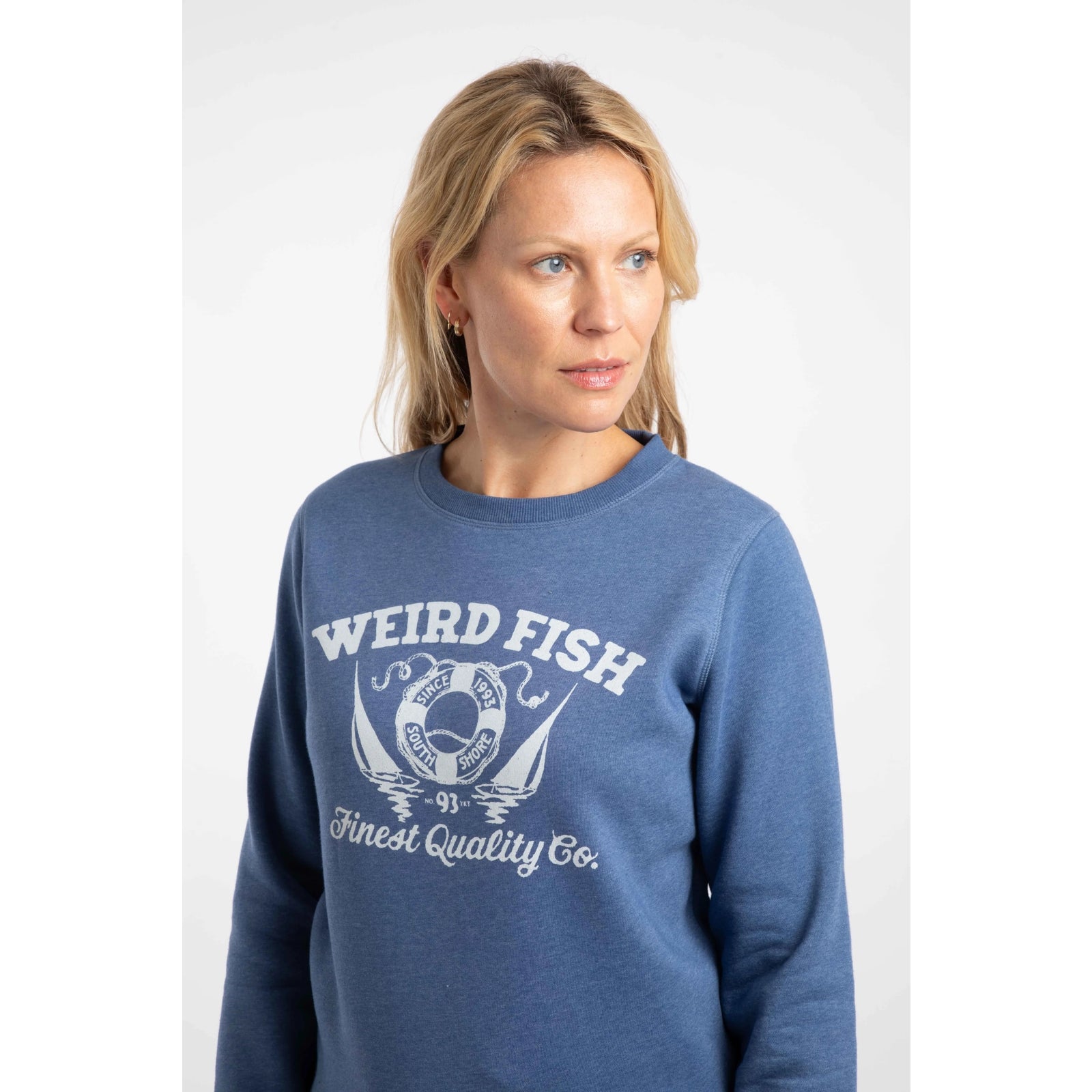 Close-up view of a woman wearing a Weird Fish Aurora crew neck sweatshirt in Faded Blue with a nautical logo print on the front.