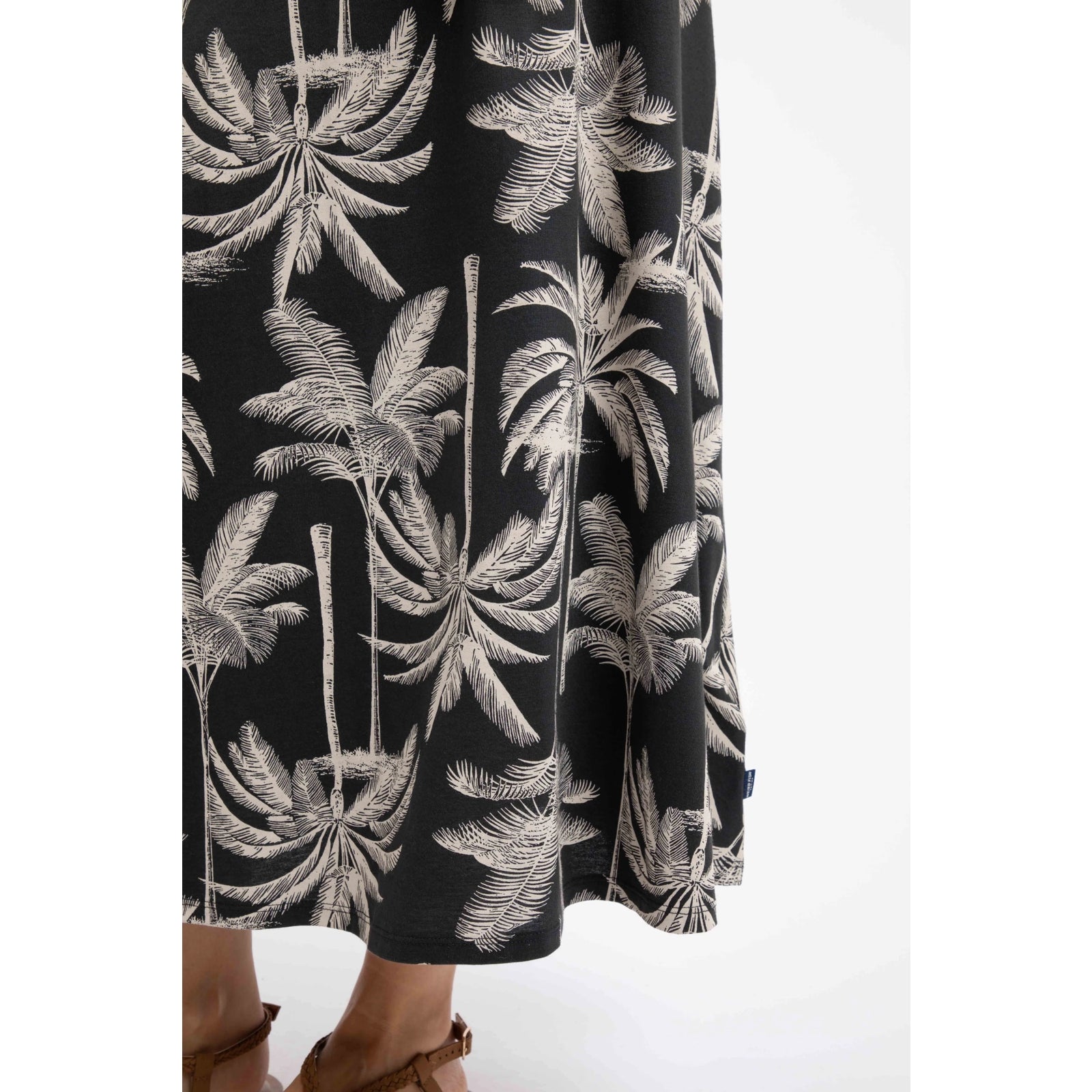 Close-up view of a woman wearing a Weird Fish Eloriah midi jersey skirt in black with a beige palm tree pattern and small logo label sewn near the hem.