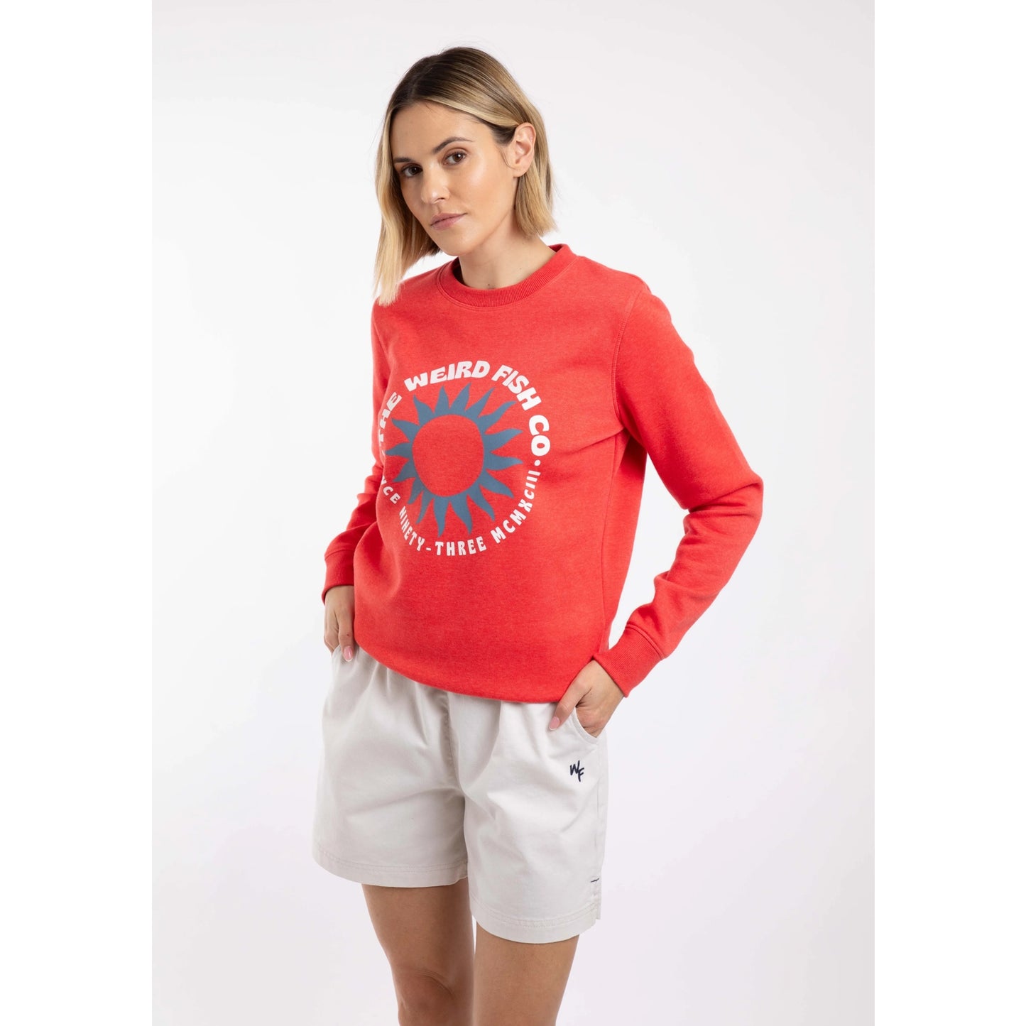Front view of a woman wearing a Weird Fish Aurora crew neck sweatshirt in a bright coral colour with bold circular logo on the front.