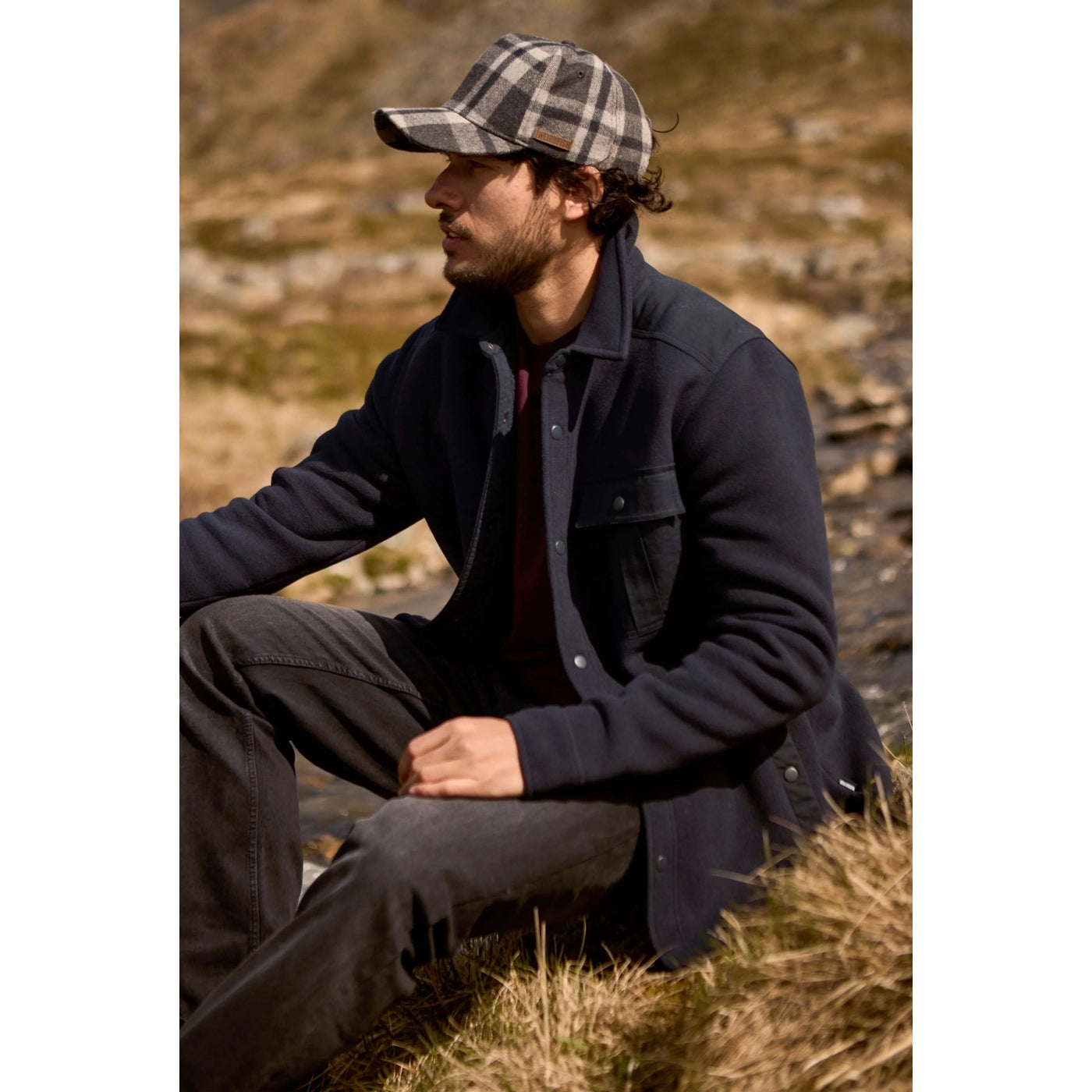 Man wearing a dark navy Weird Fish Boyd fleece jacket and plaid cap sitting in a natural setting
