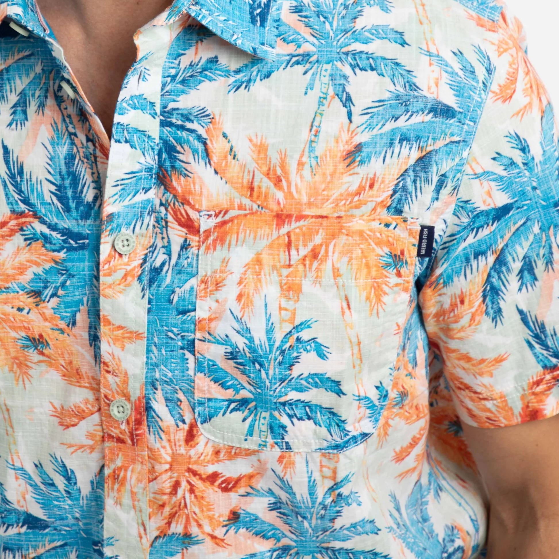 Close-up view of the chest pocket of a men's Weird Fish short sleeve Faraway shirt in Paprika orange and blue with a tropical palm tree pattern