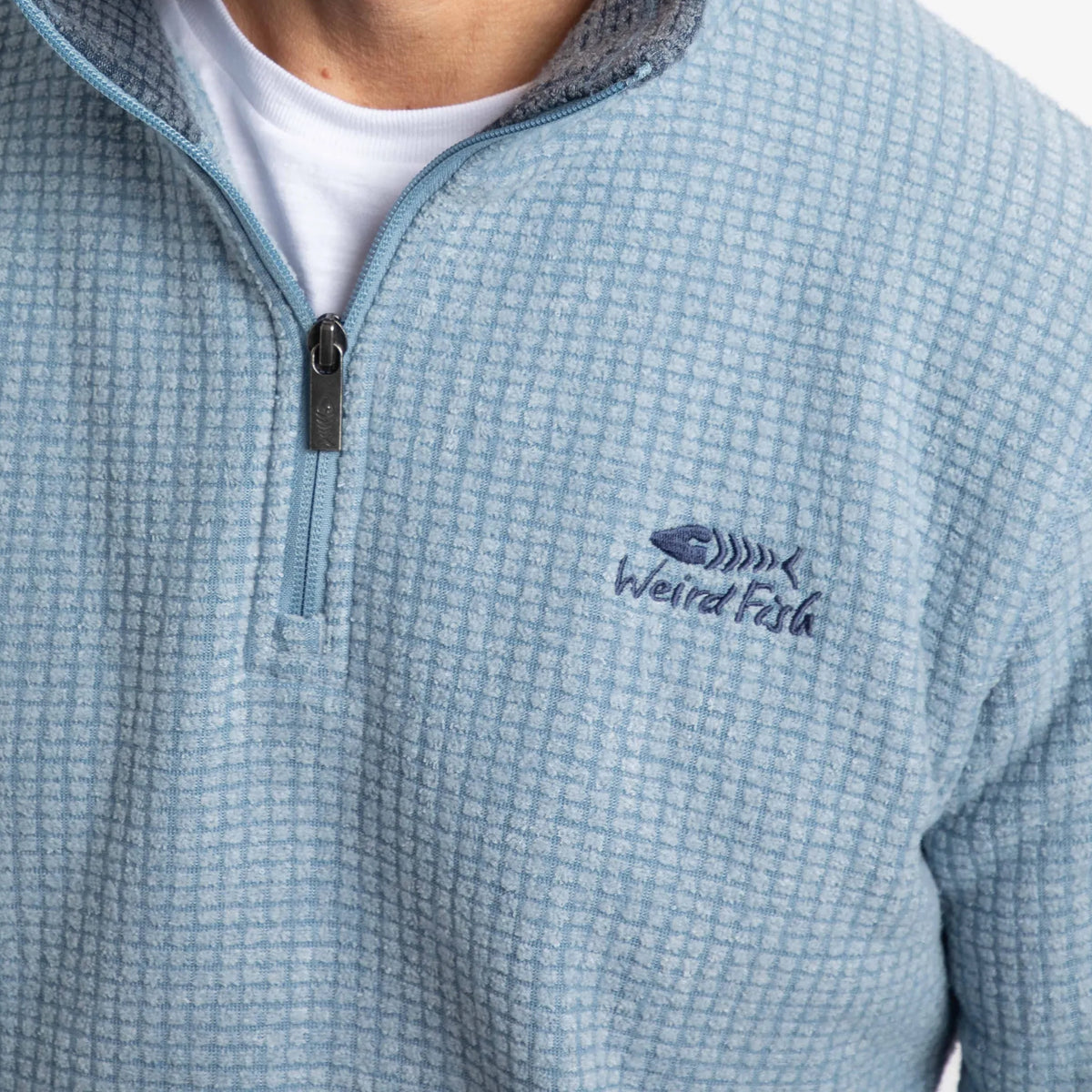 Close-up view of the 1/4 zip neckline and chest logo of a mens Weird Fish Newark grid fleece in light blue