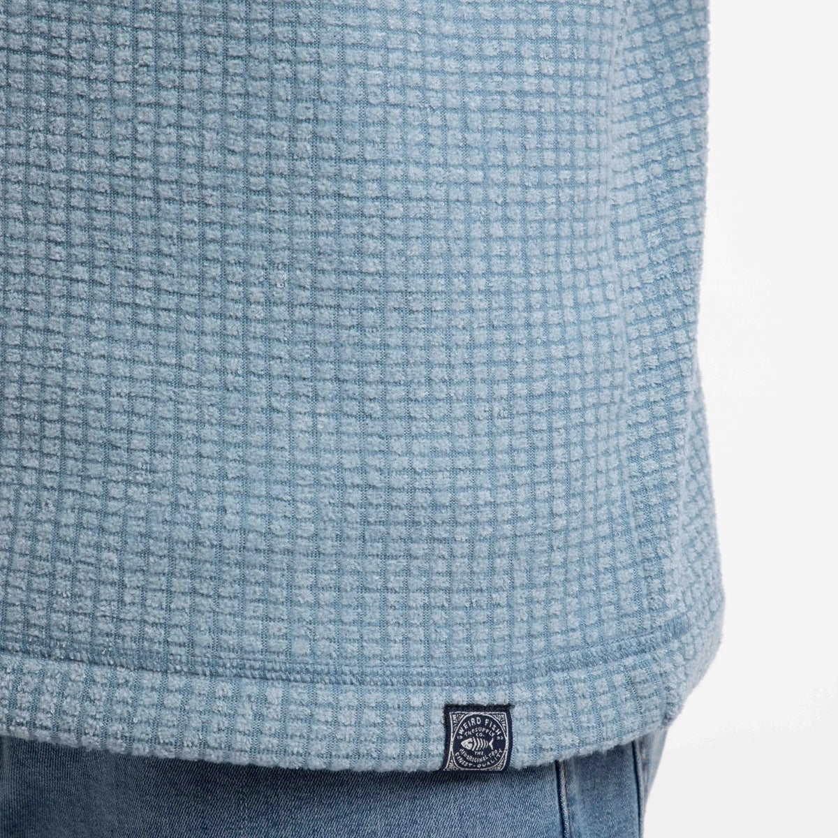Close-up view of a logo label on the hem of a mens Weird Fish Newark grid fleece in light blue