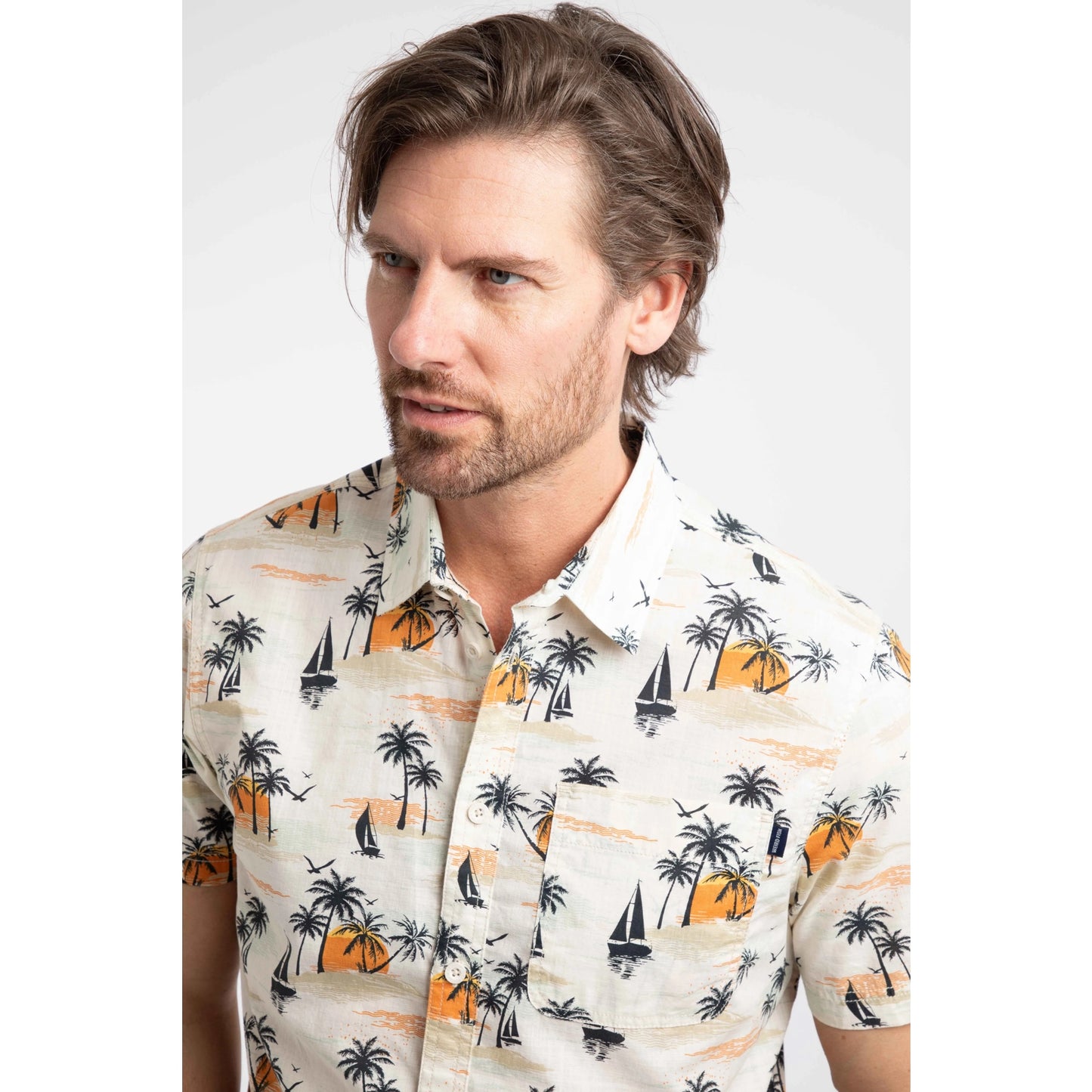 Close-up view of a man wearing a Weird Fish short sleeve Faraway shirt in Ecru with a tropical island, palm trees, sun and sailboat pattern