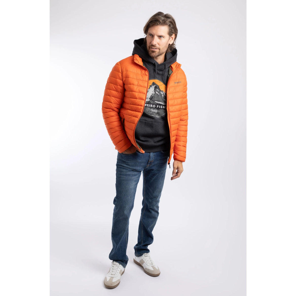 A man wearing a Weird Fish Crantock padded lightweight showerproof jacket in bright Koi Orange over a black hoodie with blue jeans.