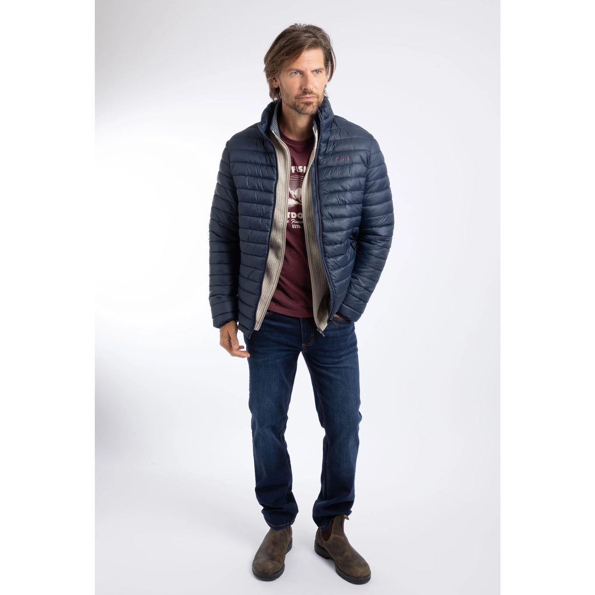 A man wearing a Weird Fish Crantock padded jacket in navy, unzipped with blue jeans and brown boots.