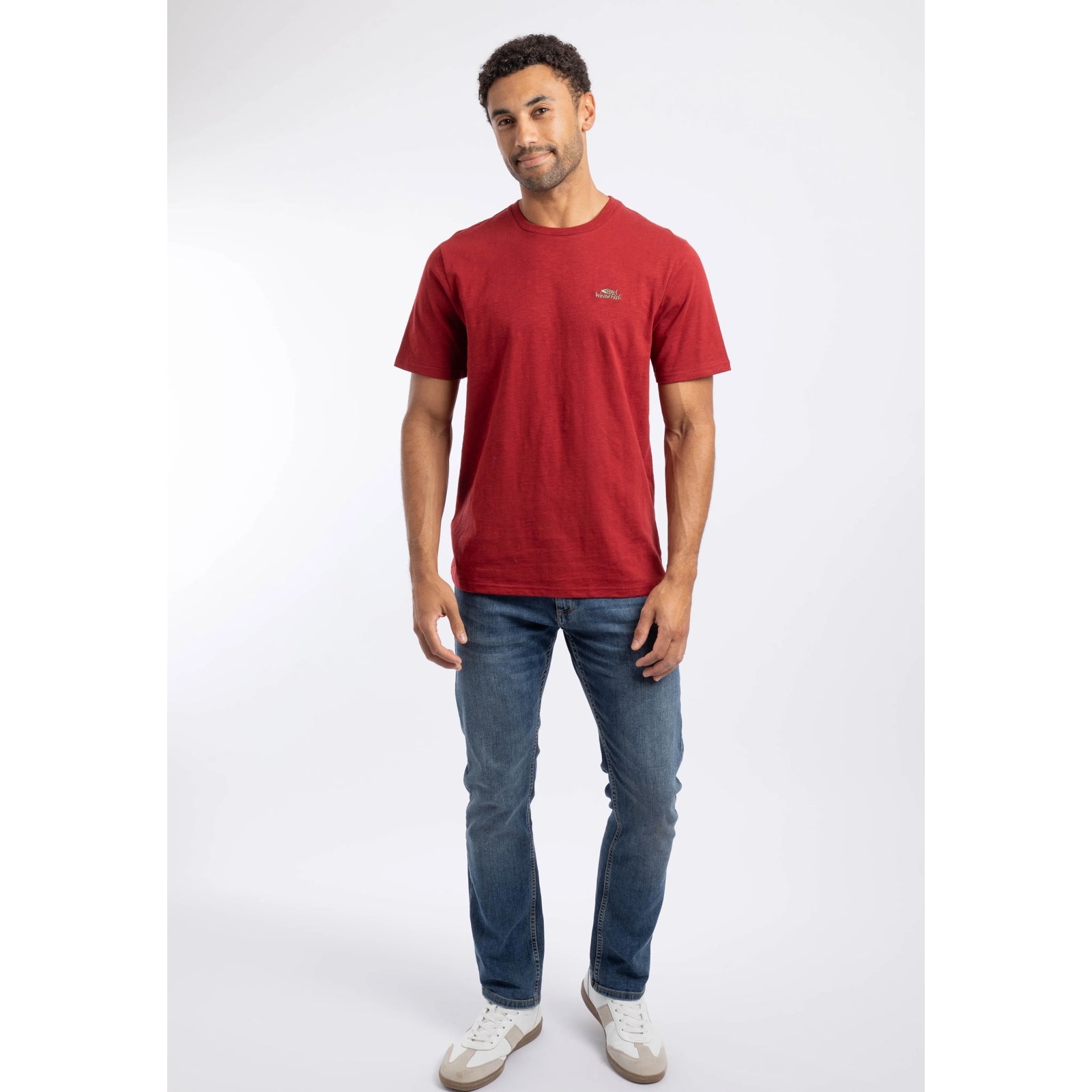 A man wearing a Weird Fish crew neck short sleeve Fished t-shirt in Barn Red with small logo on one side of the chest with blue jeans and white trainers.