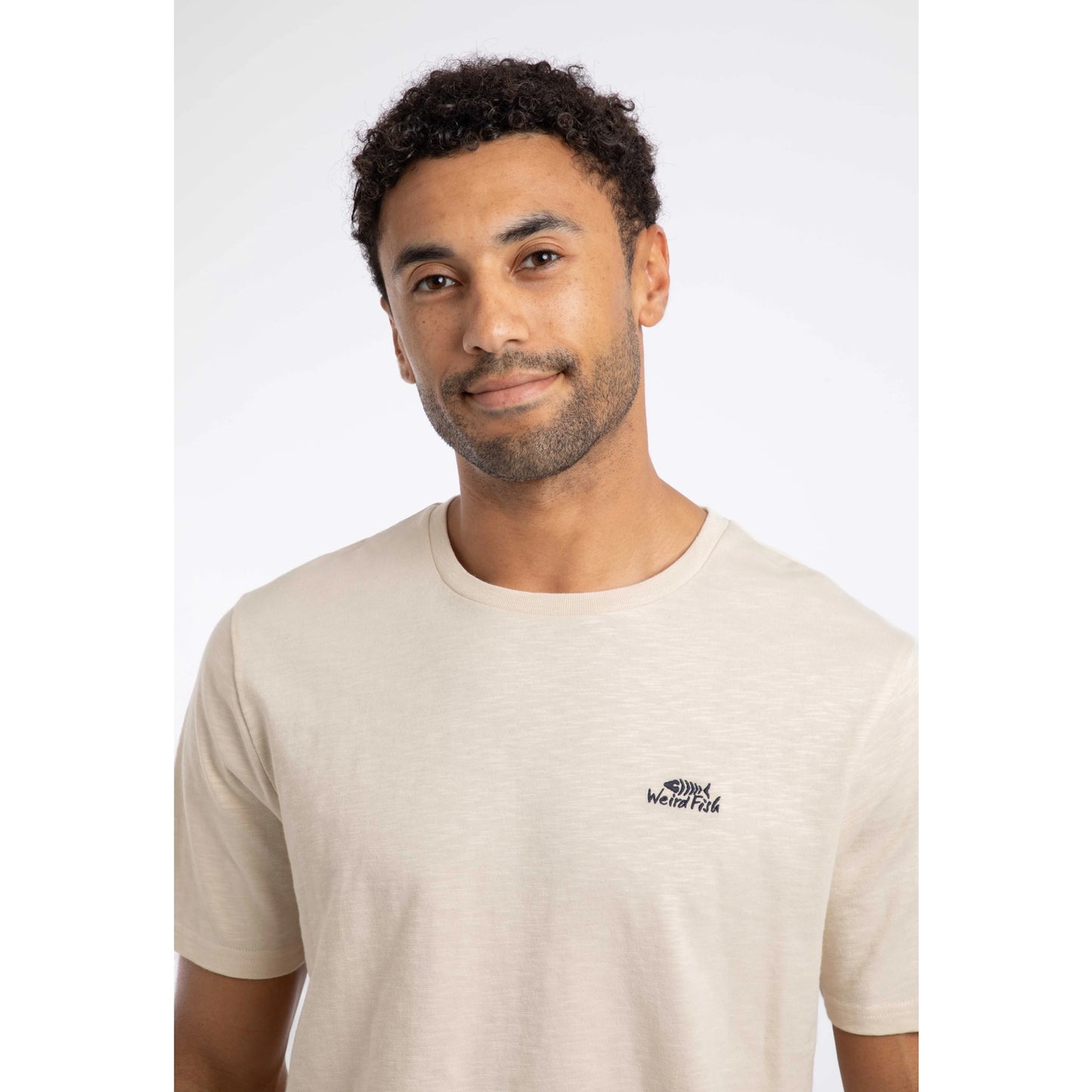 Close-up view of a man wearing a Weird Fish crew neck short sleeve Fished t-shirt in Egg Shell with small logo on one side of the chest.