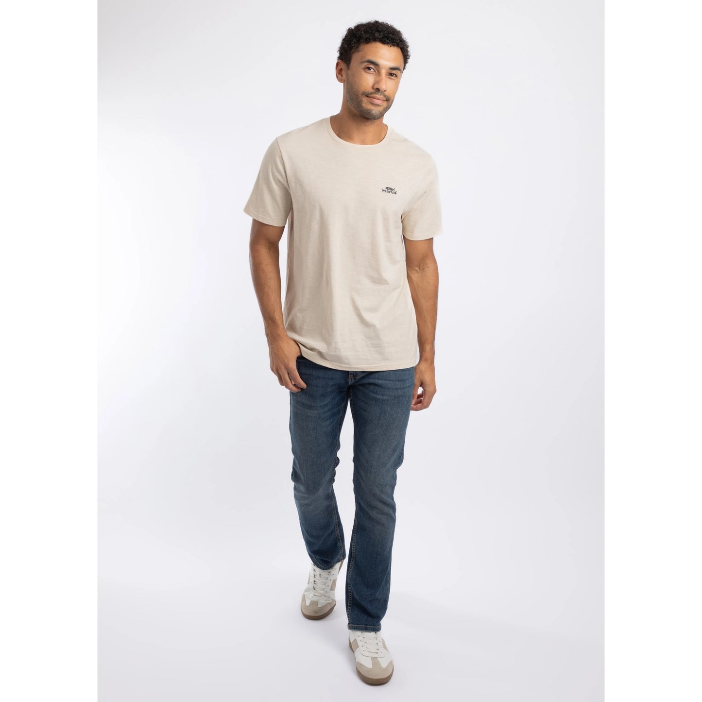 A man wearing a Weird Fish crew neck short sleeve Fished t-shirt in Egg Shell with small logo on one side of the chest with blue jeans and white trainers.