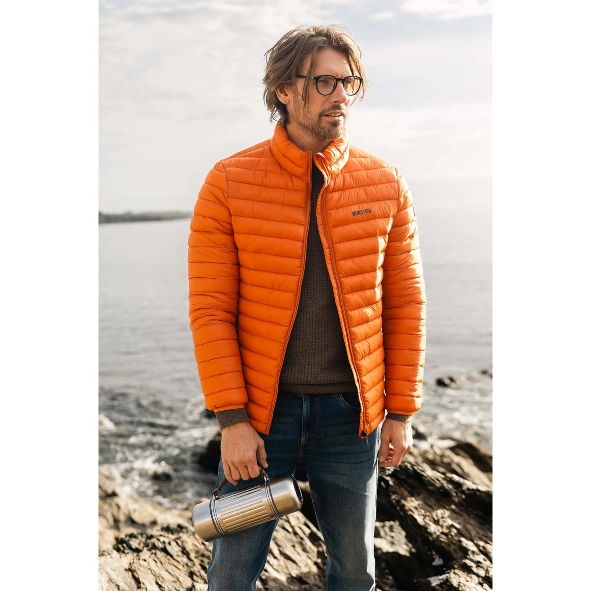 A man wearing a Weird Fish Crantock padded lightweight showerproof jacket in bright Koi Orange outdoor.