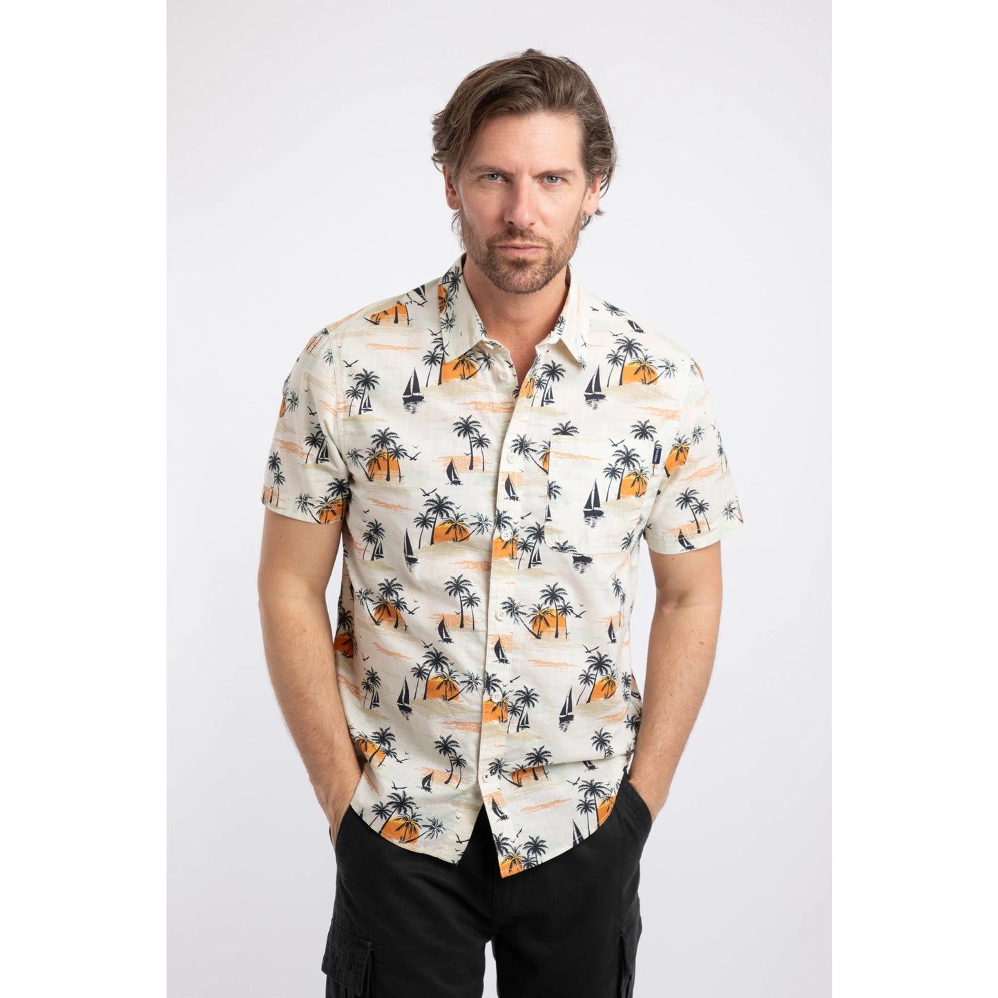 Front view of a man wearing a Weird Fish short sleeve Faraway shirt in Ecru with a tropical island, palm trees, sun and sailboat pattern