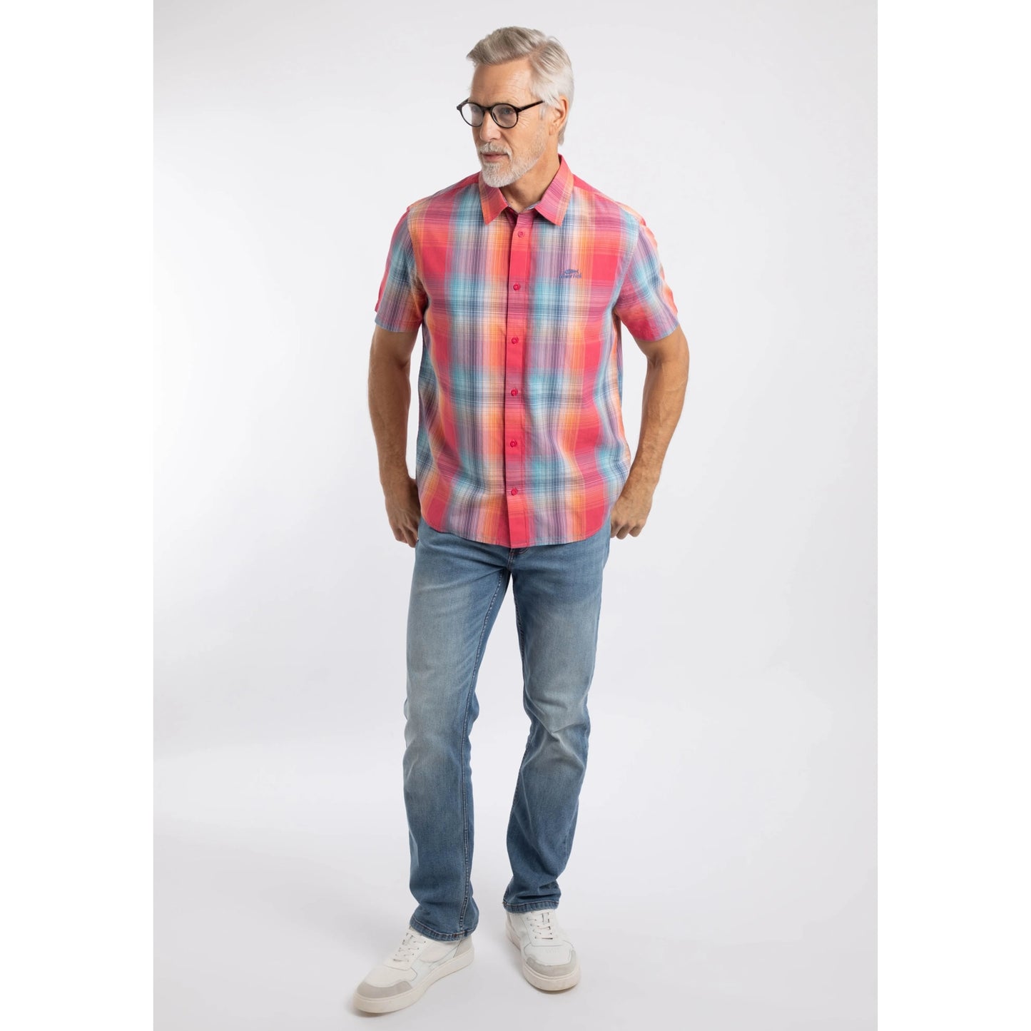 A man wearing a Weird Fish short sleeve Judd shirt in a bright Flamingo Pink, blue and orange check pattern with blue jeans and white trainers.