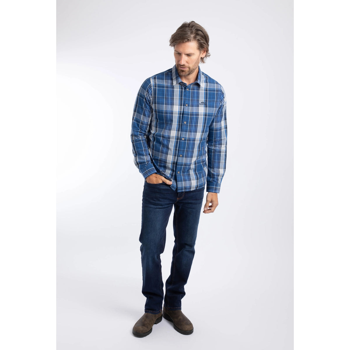 A man wearing a Weird Fish long sleeve Lando shirt in a blue, navy and white check pattern, paired with blue jeans and brown boots.