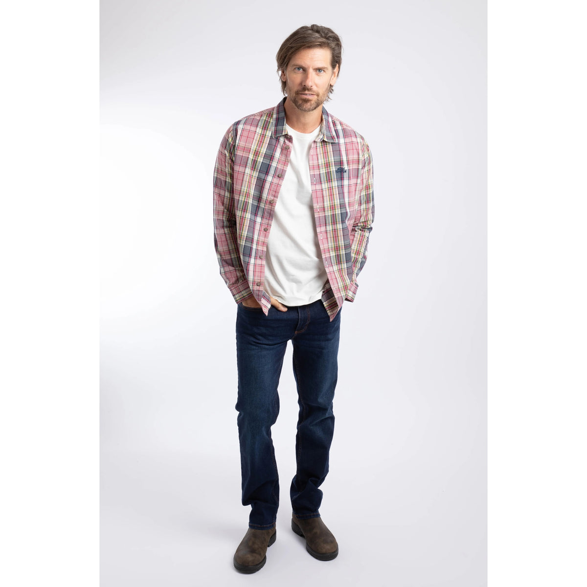 A man wearing a Weird Fish long sleeve Lando shirt, in a crushed berry, navy, yellow and white check pattern paired with blue jeans and brown boots.