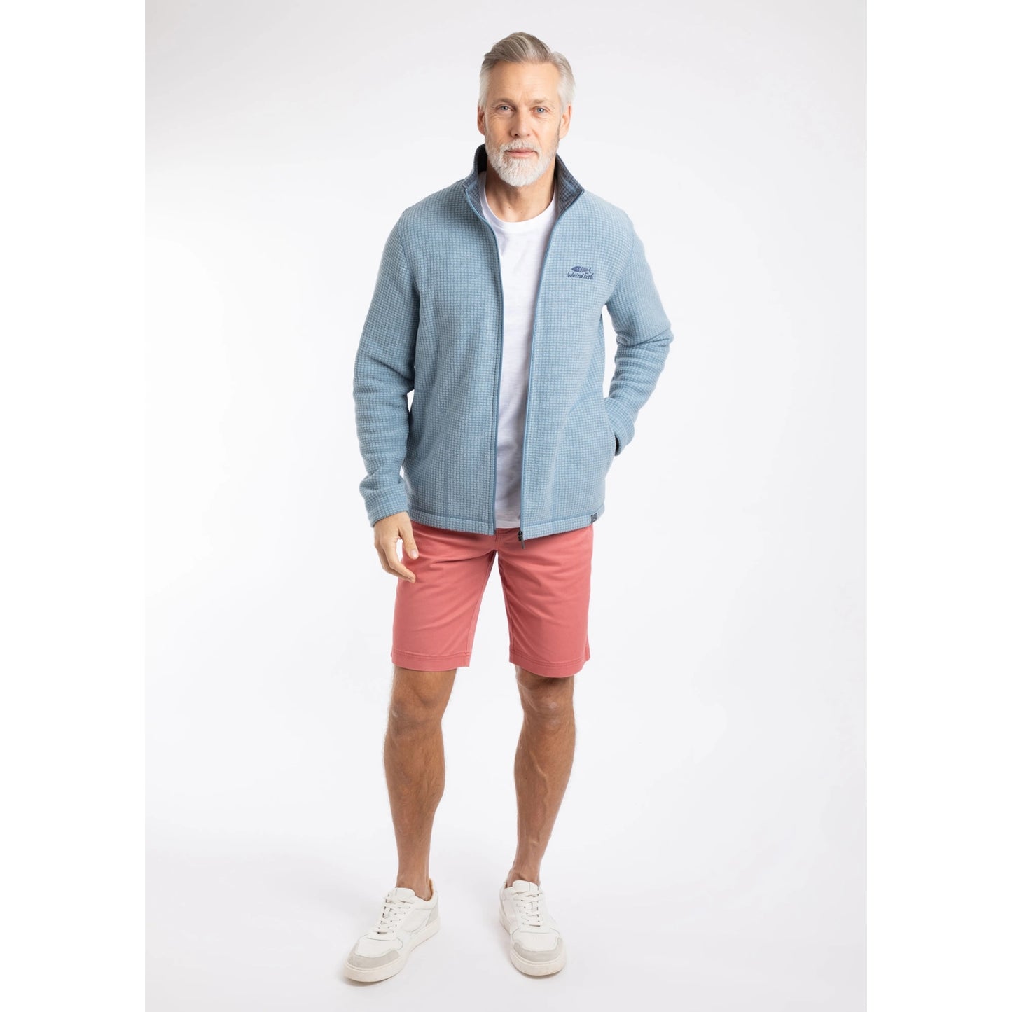 Front view of a man wearing a Weird Fish full zip Shires grid textured fleece in Light Blue unzipped with a white t-shirt, contrasting navy neckline and small chest logo with pink shorts
