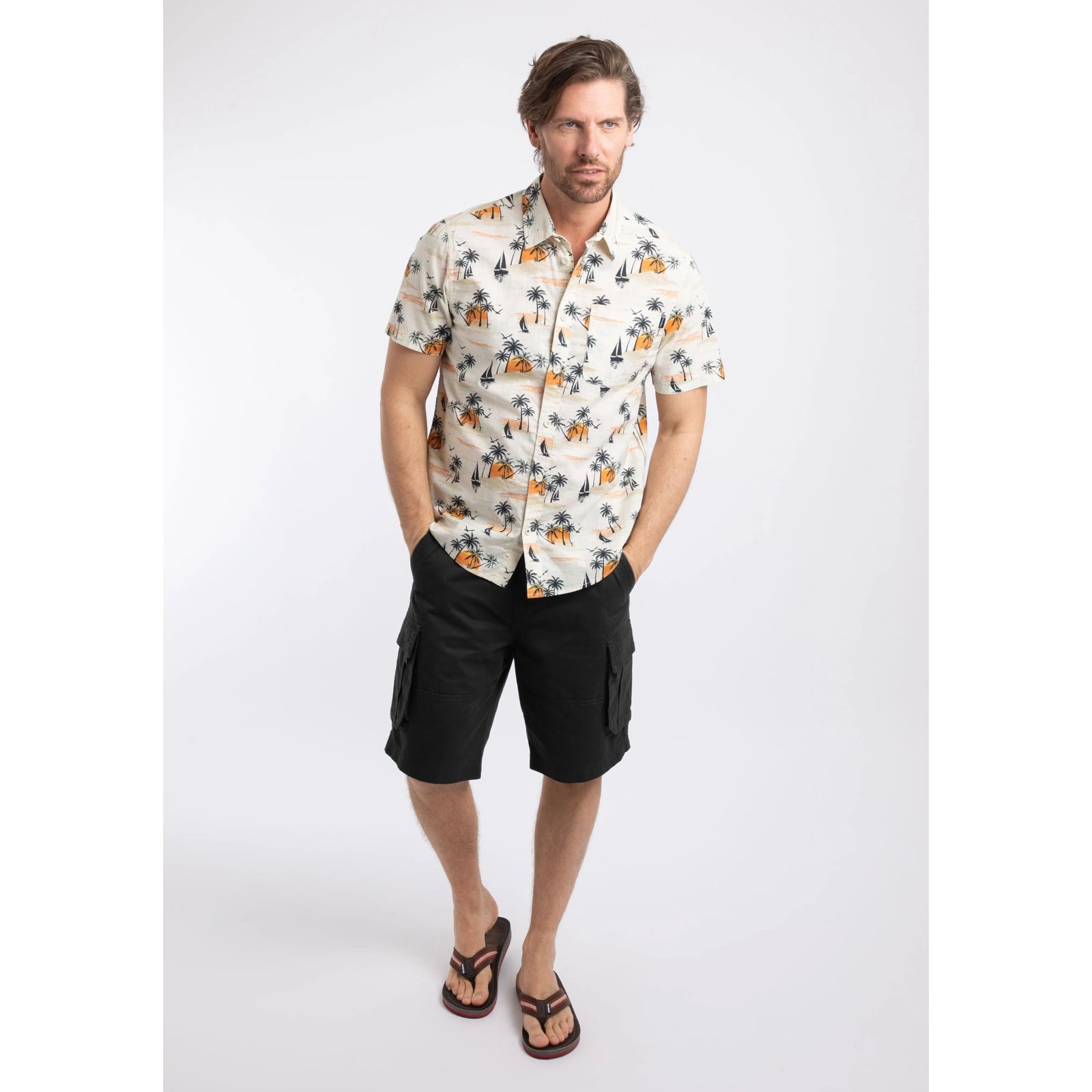 Front view of a man wearing a Weird Fish short sleeve Faraway shirt in Ecru with a tropical island, palm trees, sun and sailboat pattern with navy shorts and flip flops.
